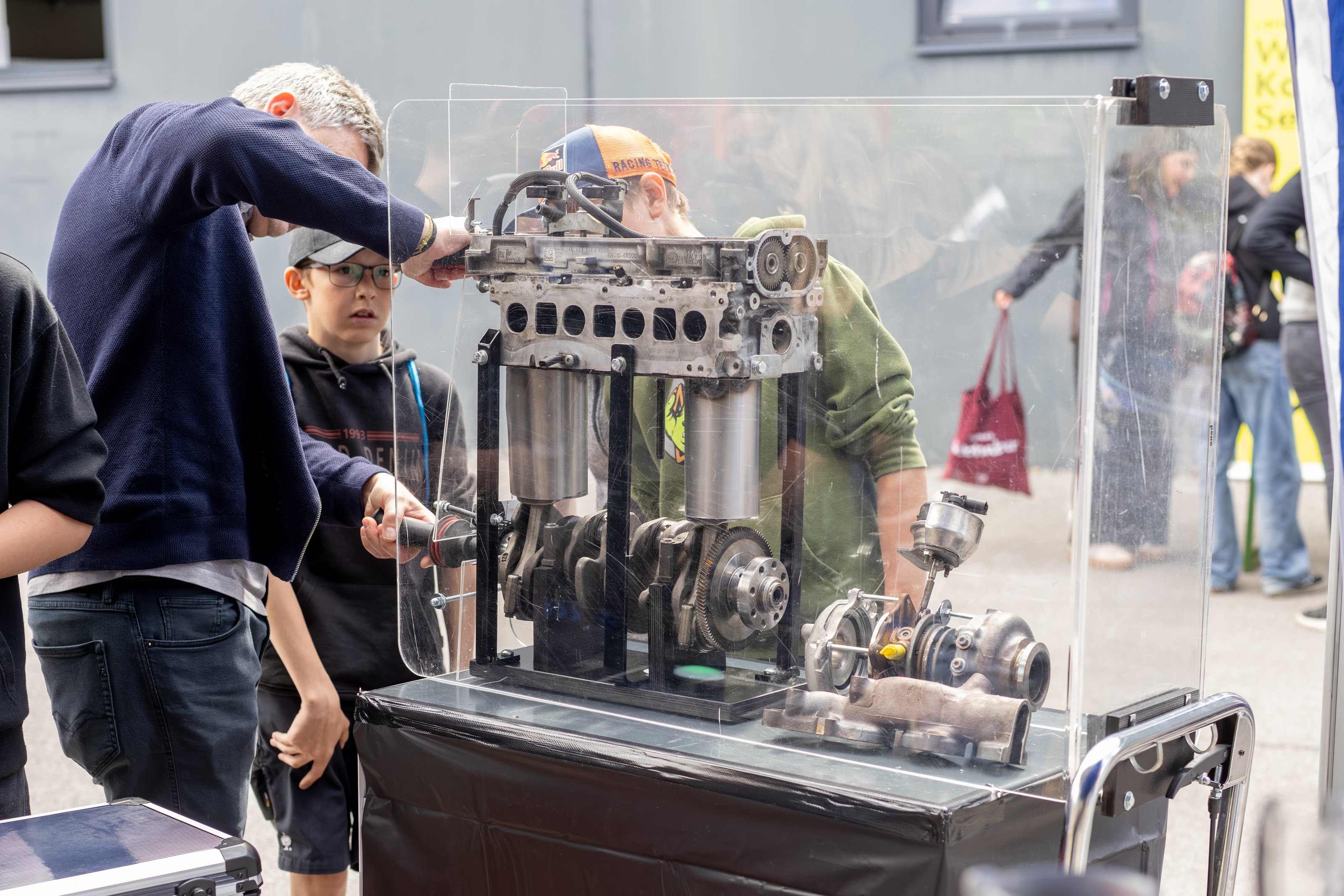 Mehrere Personen betrachten bei einer Mitmachstation einen ge&ouml;ffneten Motor hinter einer transparenten Schutzwand. Eine Person erkl&auml;rt technische Details.