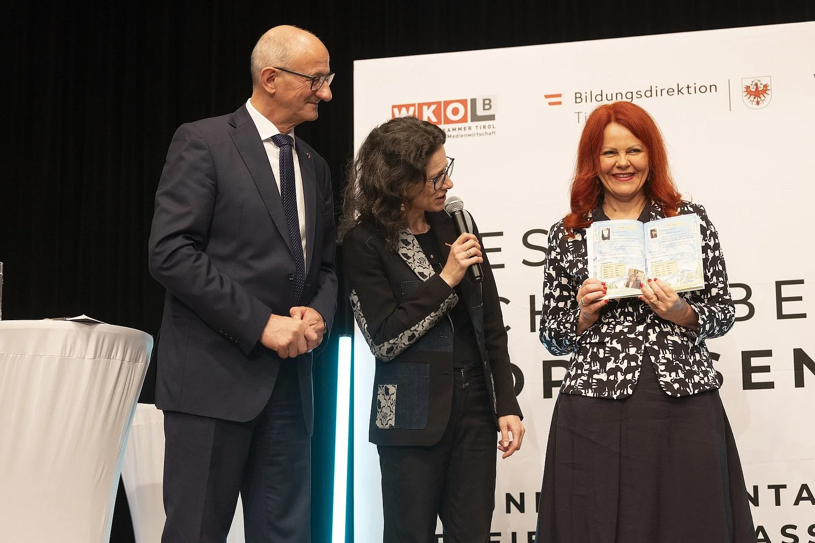 Eine Person steht auf einer B&uuml;hne und h&auml;lt ein ge&ouml;ffnetes Buch in den H&auml;nden, das sie dem Publikum zeigt. Neben ihr stehen zwei weitere Personen, eine davon spricht in ein Mikrofon. Im Hintergrund sind Logos der Veranstalter zu sehen. Die Szene zeigt die Pr&auml;sentation des Tiroler Vorlesebuchs.
