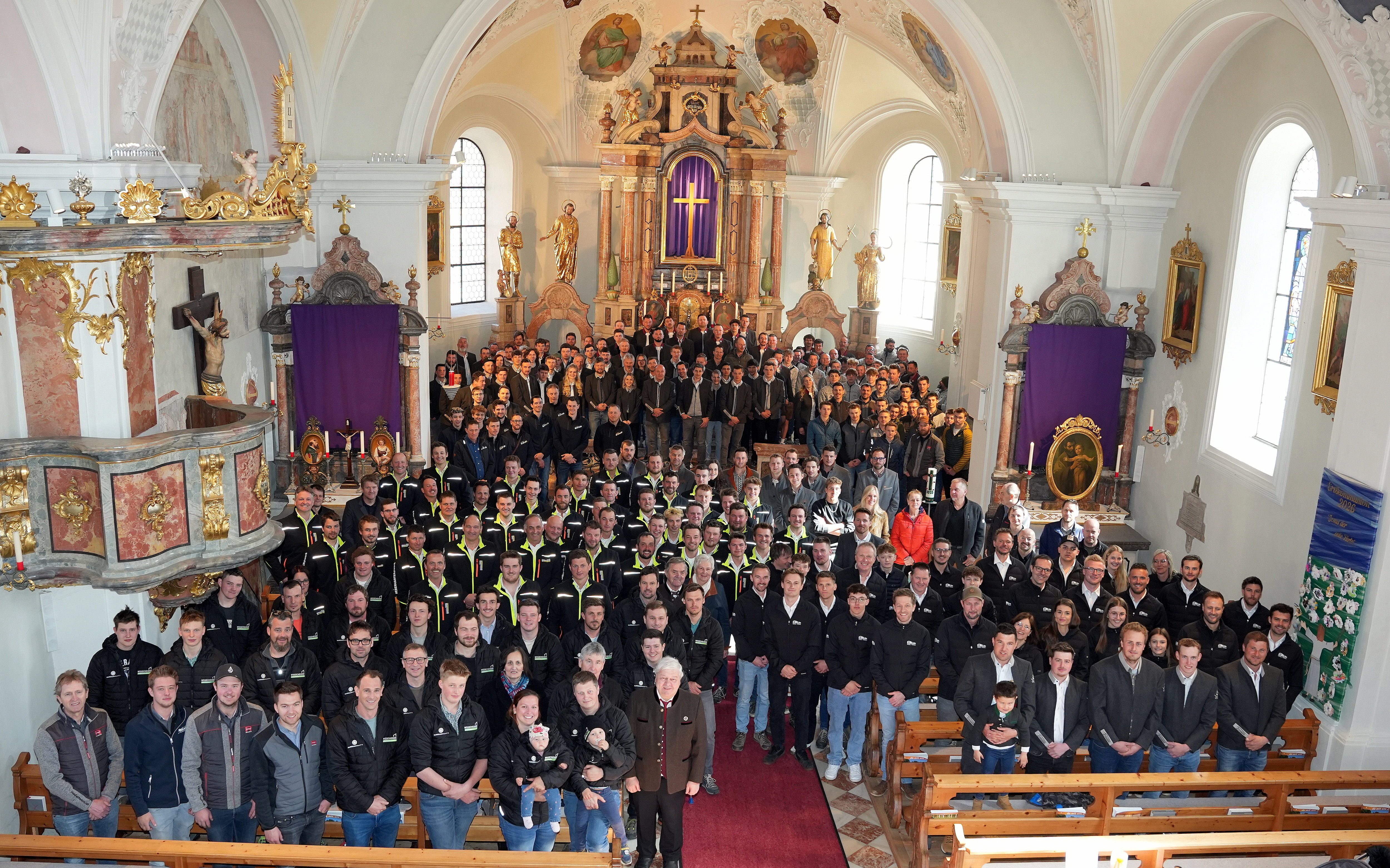 In der hellen Pfarrkirche M&uuml;nster steht eine gro&szlig;e Gruppe von rund 300 Zimmerinnen und Zimmerern sowie Branchenvertreter:innen dicht gedr&auml;ngt vor dem Altar f&uuml;r ein Gruppenfoto. Die Teilnehmenden tragen &uuml;berwiegend Arbeitskleidung oder dunkle Jacken mit Firmenlogos. Im Hintergrund ist der reich verzierte Kirchenaltar mit goldenen Elementen und religi&ouml;sen Darstellungen zu sehen. Die Aufnahme vermittelt den starken Zusammenhalt innerhalb der Holzbaubranche und die Bedeutung des gemeinsamen Josefi-Tages.