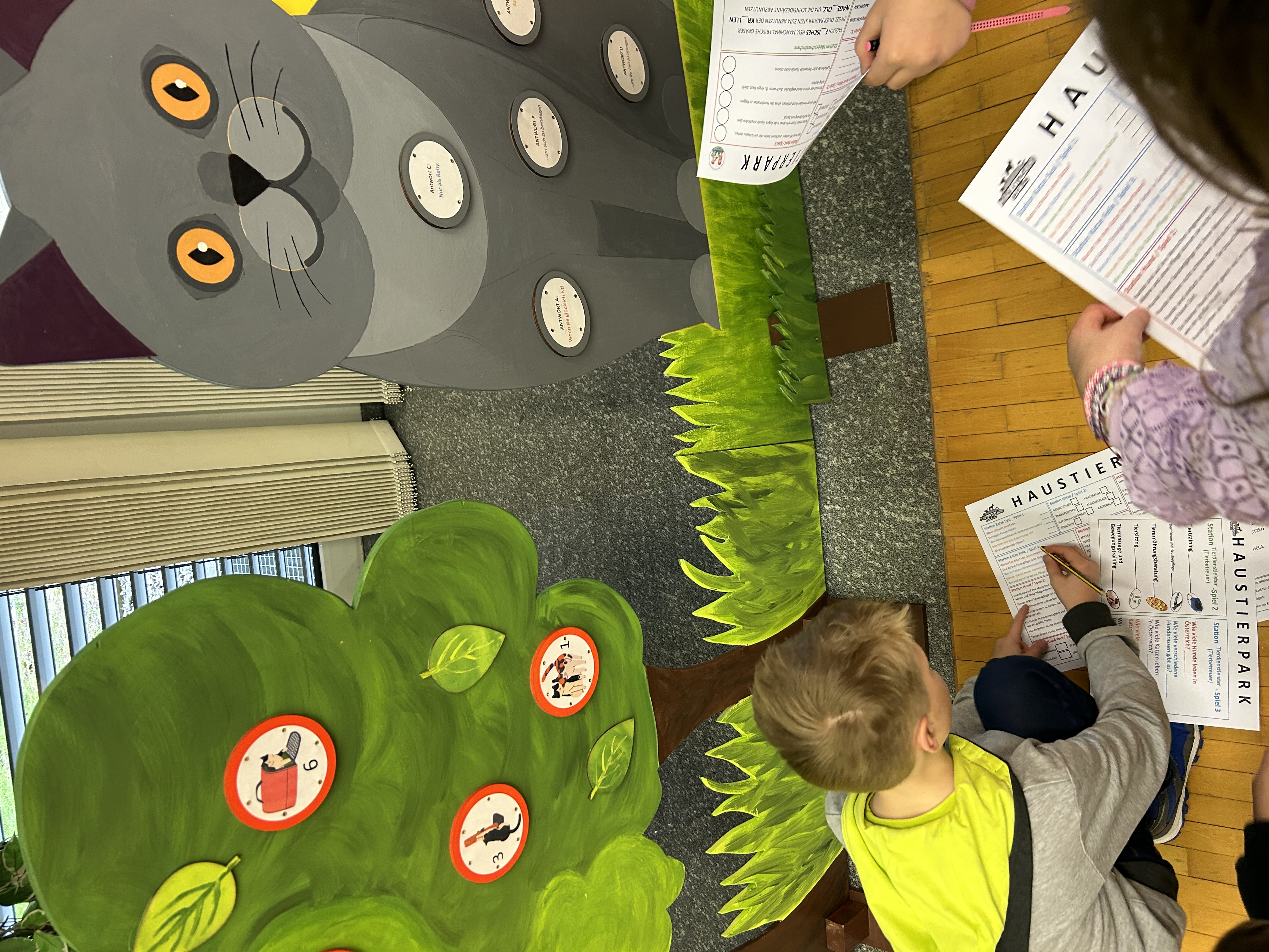Kinder bearbeiten ein Arbeitsblatt vor einer Station mit einer gro&szlig;en, grauen Katzenfigur und einem gr&uuml;nen Baum mit Symbolen. Die Kinder stehen oder knien am Boden und schreiben auf ihre Unterlagen.