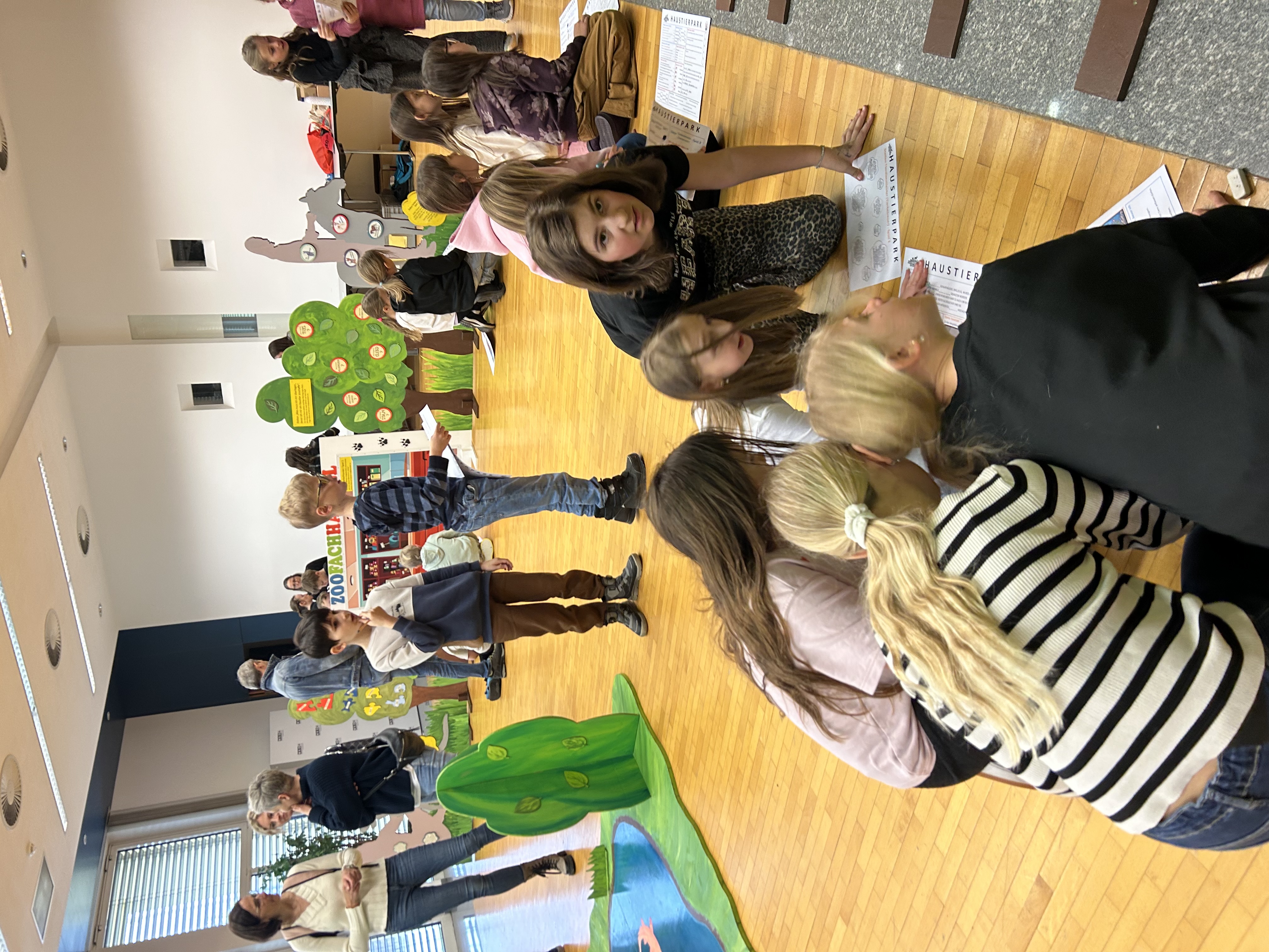 Mehrere Kinder bewegen sich in einem Raum zwischen verschiedenen Stationen mit Tiermotiven. Einige knien am Boden und bearbeiten Arbeitsbl&auml;tter, andere gehen zwischen den aufgebauten Elementen umher. Im Hintergrund stehen auch Erwachsene und beobachten das Geschehen.