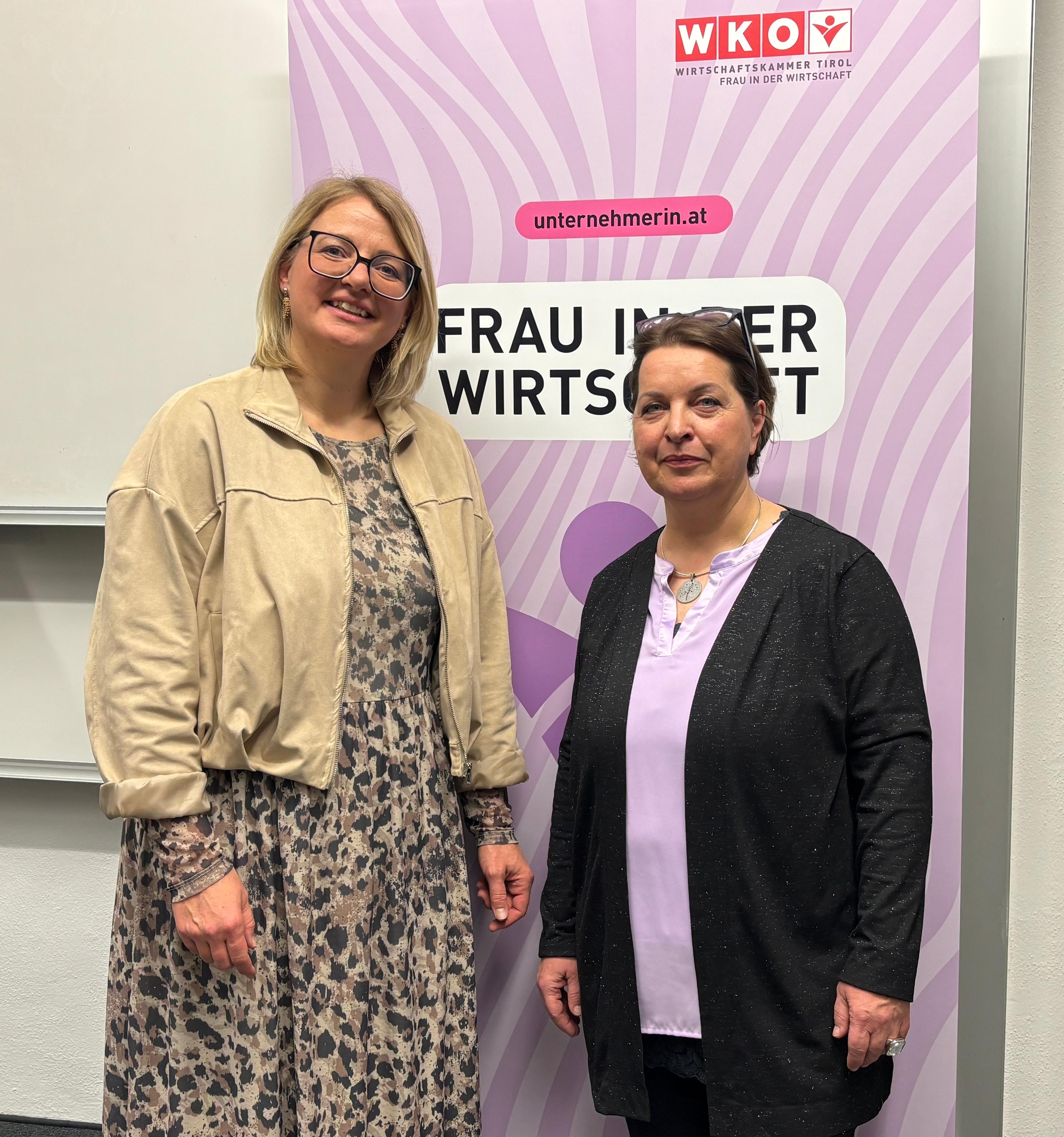 Zwei Frauen stehen nebeneinander vor einem Roll-up der Wirtschaftskammer Tirol &ndash; Frau in der Wirtschaft. Die Frau links tr&auml;gt ein gemustertes Kleid mit heller Jacke und l&auml;chelt in die Kamera. Die Frau rechts tr&auml;gt ein dunkles Outfit mit lila Oberteil und blickt ebenfalls in die Kamera. Im Hintergrund ist das Logo der Wirtschaftskammer Tirol sowie der Schriftzug &bdquo;Frau in der Wirtschaft&ldquo; und die Website unternehmerin.at zu sehen. Die Aufnahme entstand in einem Innenraum bei einer Veranstaltung.