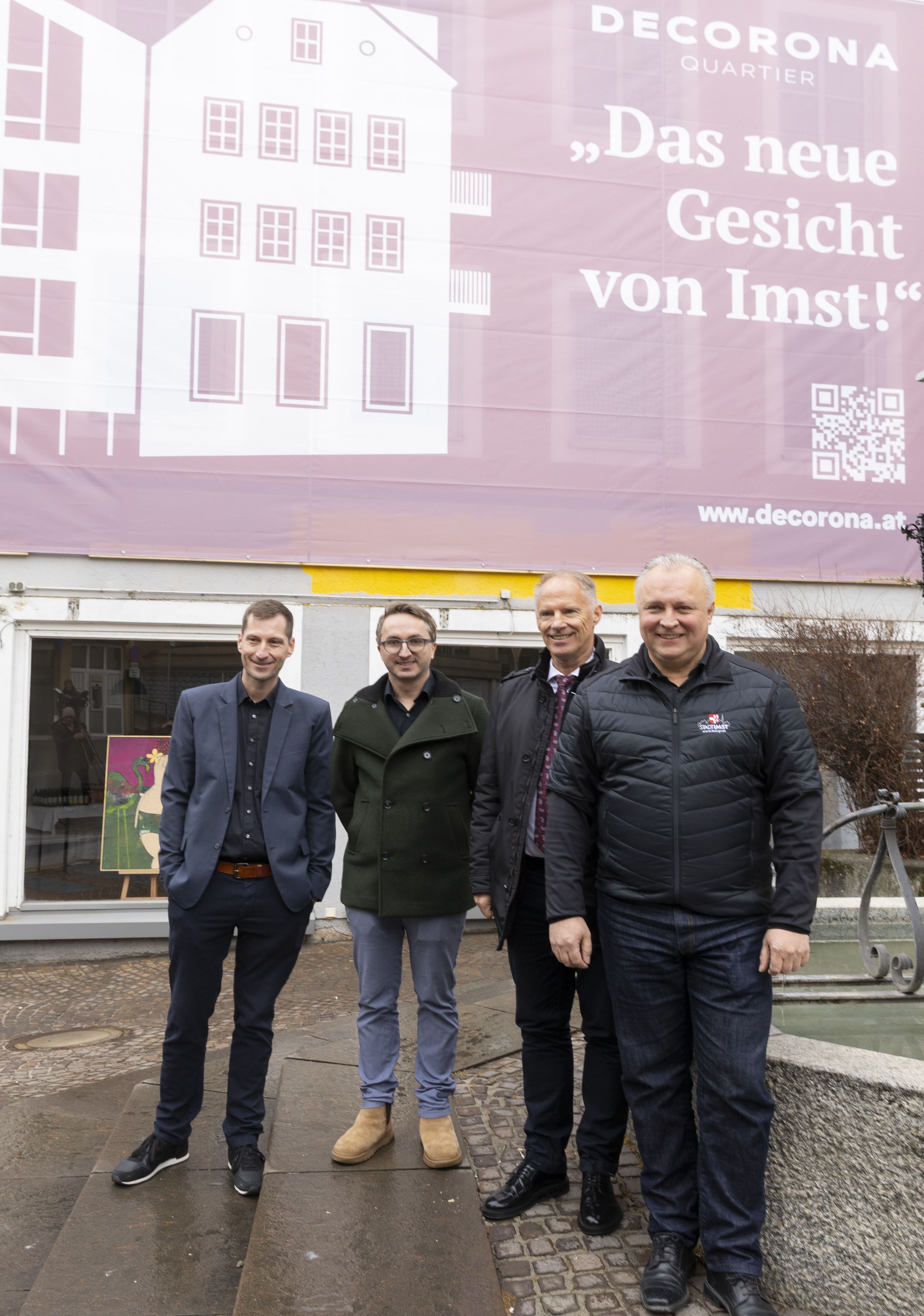 Vier Projektverantwortliche stehen vor einer gro&szlig;fl&auml;chigen Baustellenplane mit der Aufschrift &bdquo;Decorona Quartier &ndash; Das neue Gesicht von Imst&ldquo;. Sie posieren vor einem Geb&auml;ude im Bestand, im Vordergrund ein Brunnen. Die Szene dokumentiert den Projektstart und zeigt Vertreter aus Projektentwicklung, Architektur, Finanzierung und Stadtpolitik.