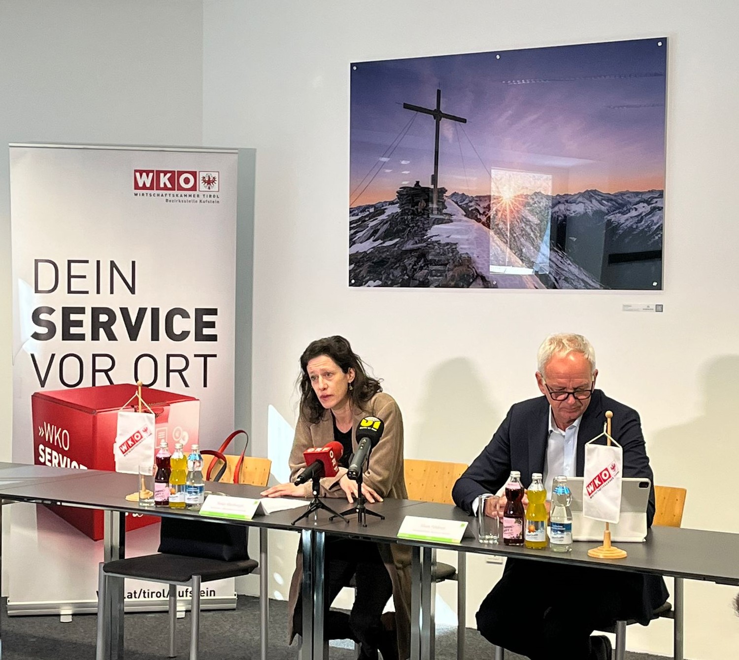 Eine Person sitzt an einem Tisch und spricht in Mikrofone. Neben ihr sitzt eine andere Person und blickt nach unten auf Unterlagen