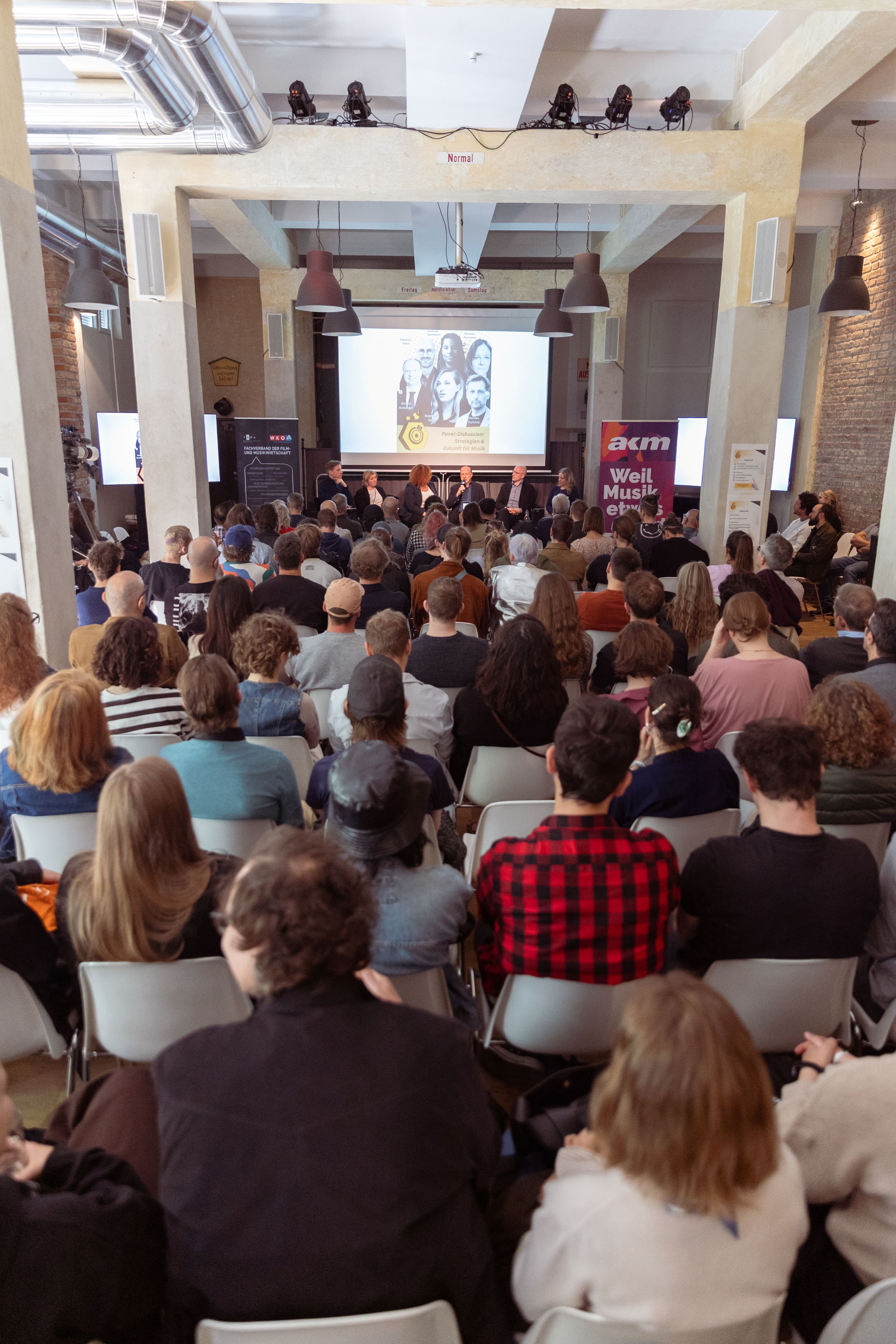 Publikum bei Podiumsdiskussion vor B&uuml;hne mit Pr&auml;sentationsfolie und Veranstaltungsbannern