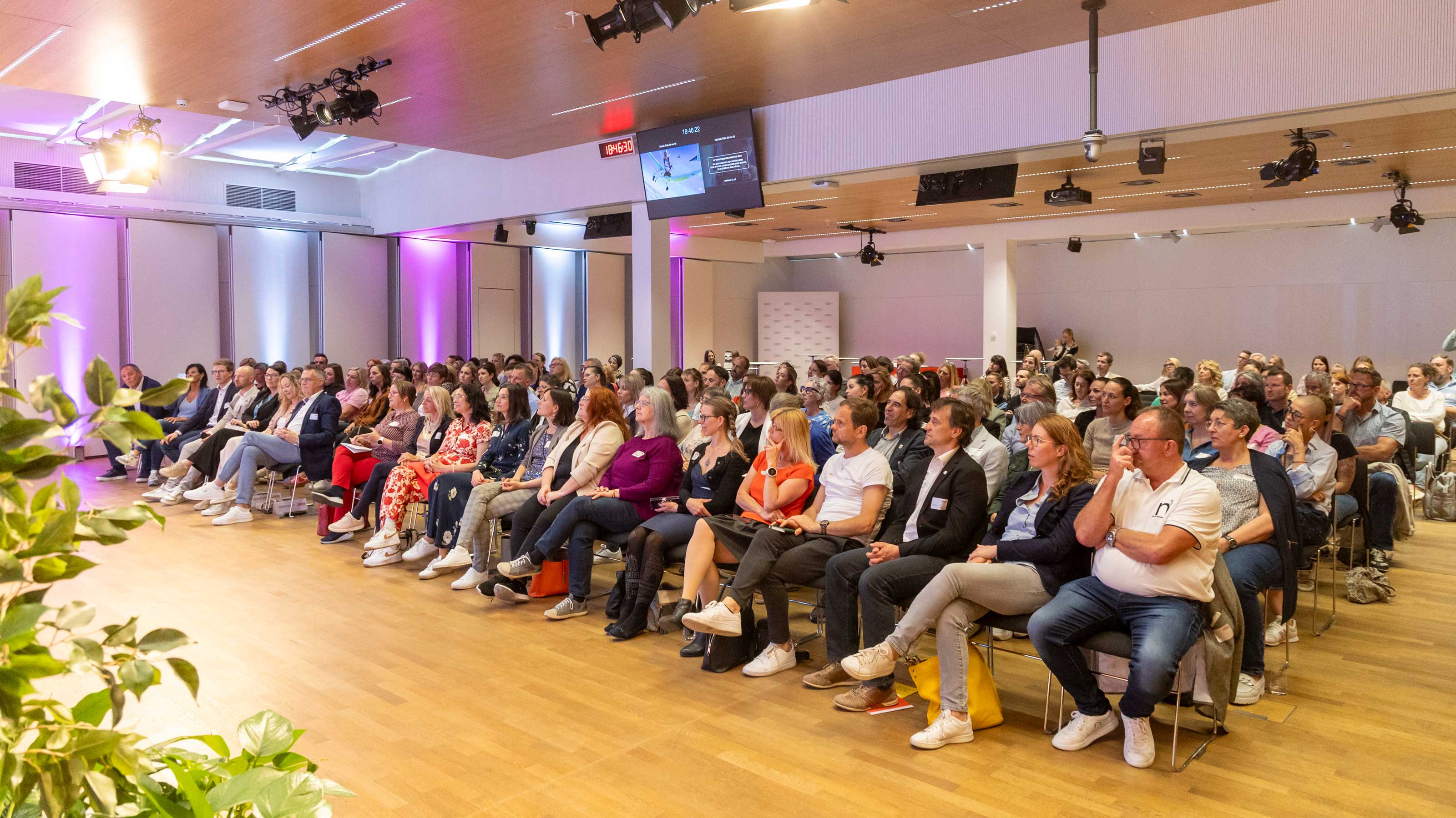 Blick in einen Veranstaltungssaal, man sieht das Publikum, das in Reihen sitzt und den Blick zum Podium gerichtet hat