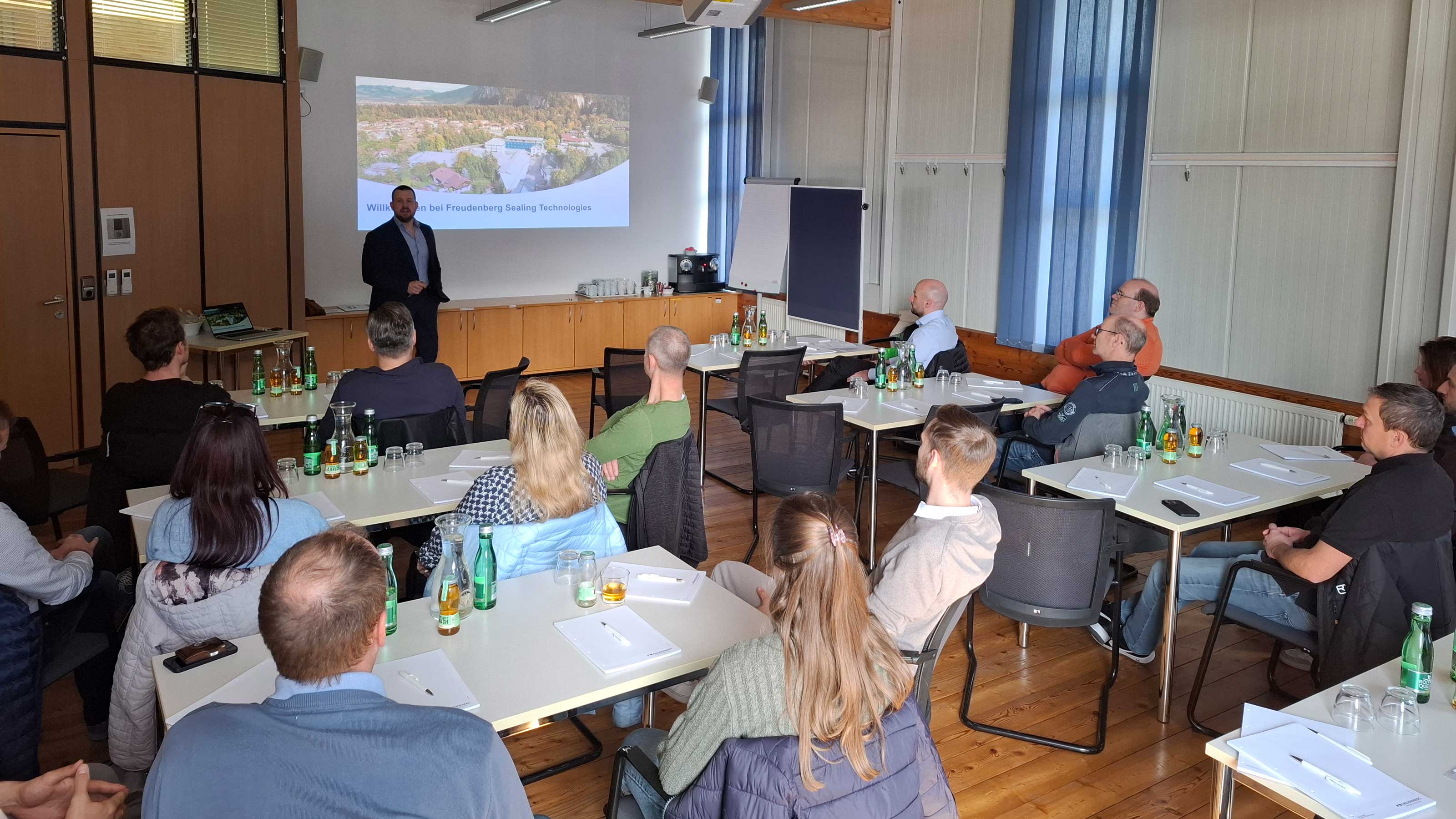 Raum mit mehreren Personen, die an Tischen sitzen, vorne steht ein Referent und h&auml;lt eine Pr&auml;sentation