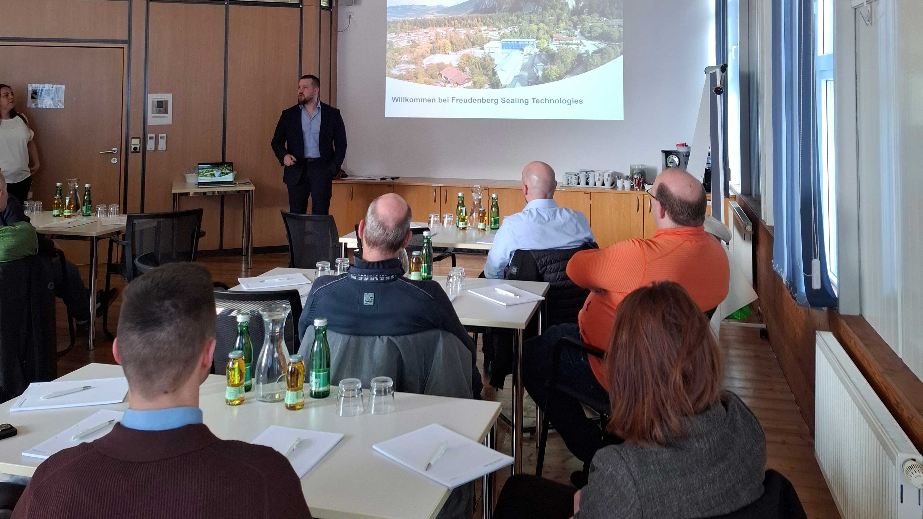 Raum mit mehreren Personen, die an Tischen sitzen, vorne steht ein Referent und h&auml;lt eine Pr&auml;sentation