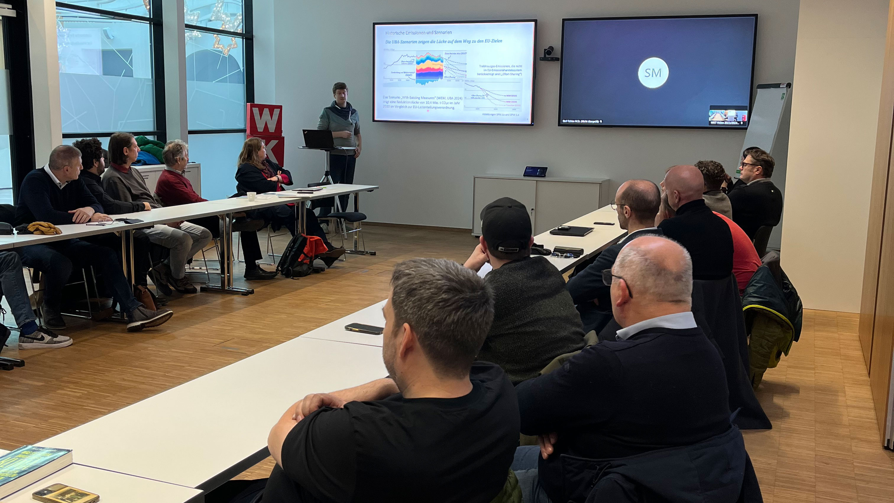 Personen sitzen in einem Seminarraum und h&ouml;ren dem Vortrag des Referenten zu, der vorne steht und seine Folien pr&auml;sentiert