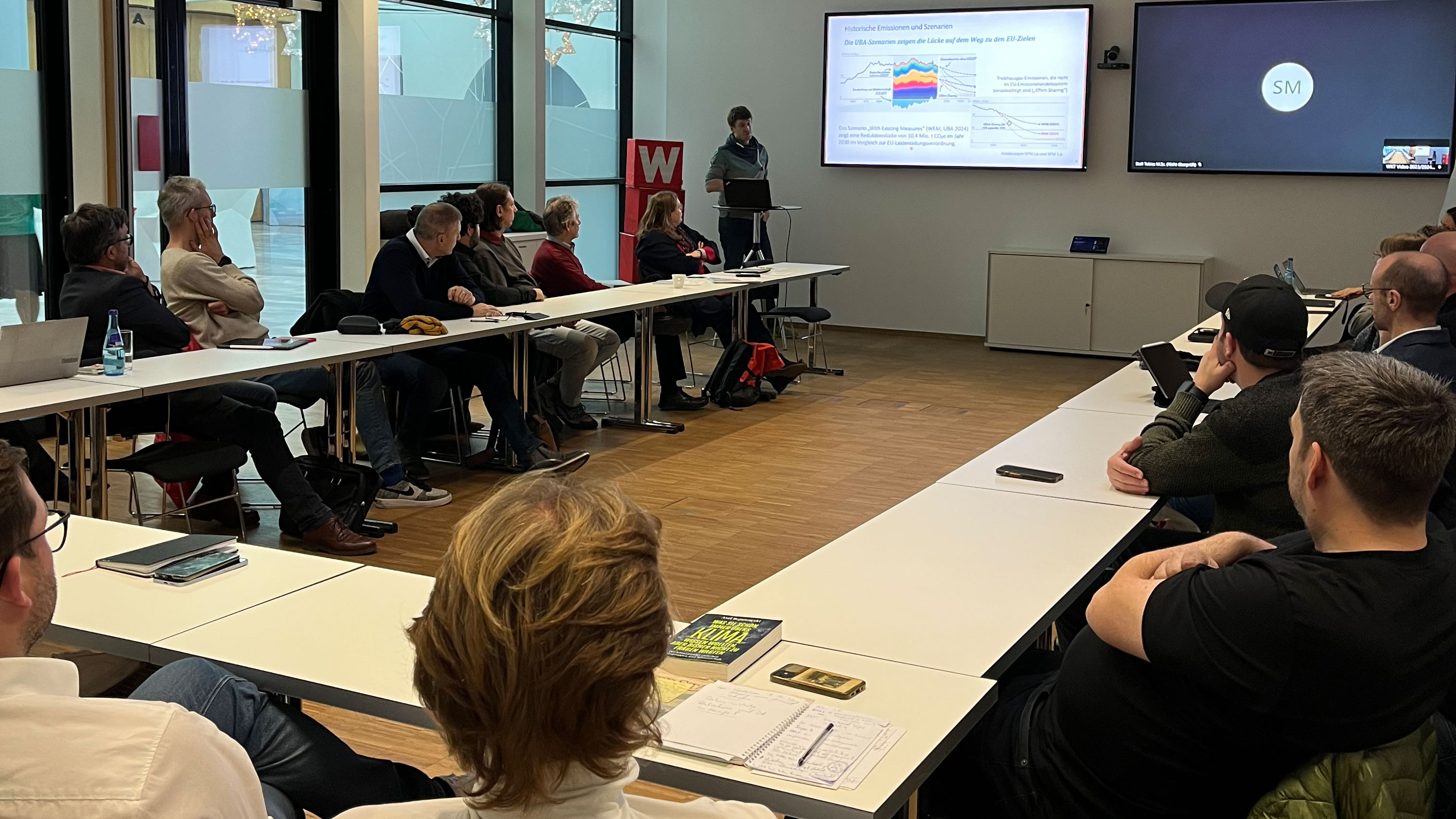 Personen sitzen in einem Seminarraum und h&ouml;ren dem Vortrag des Referenten zu, der vorne steht und seine Folien pr&auml;sentiert