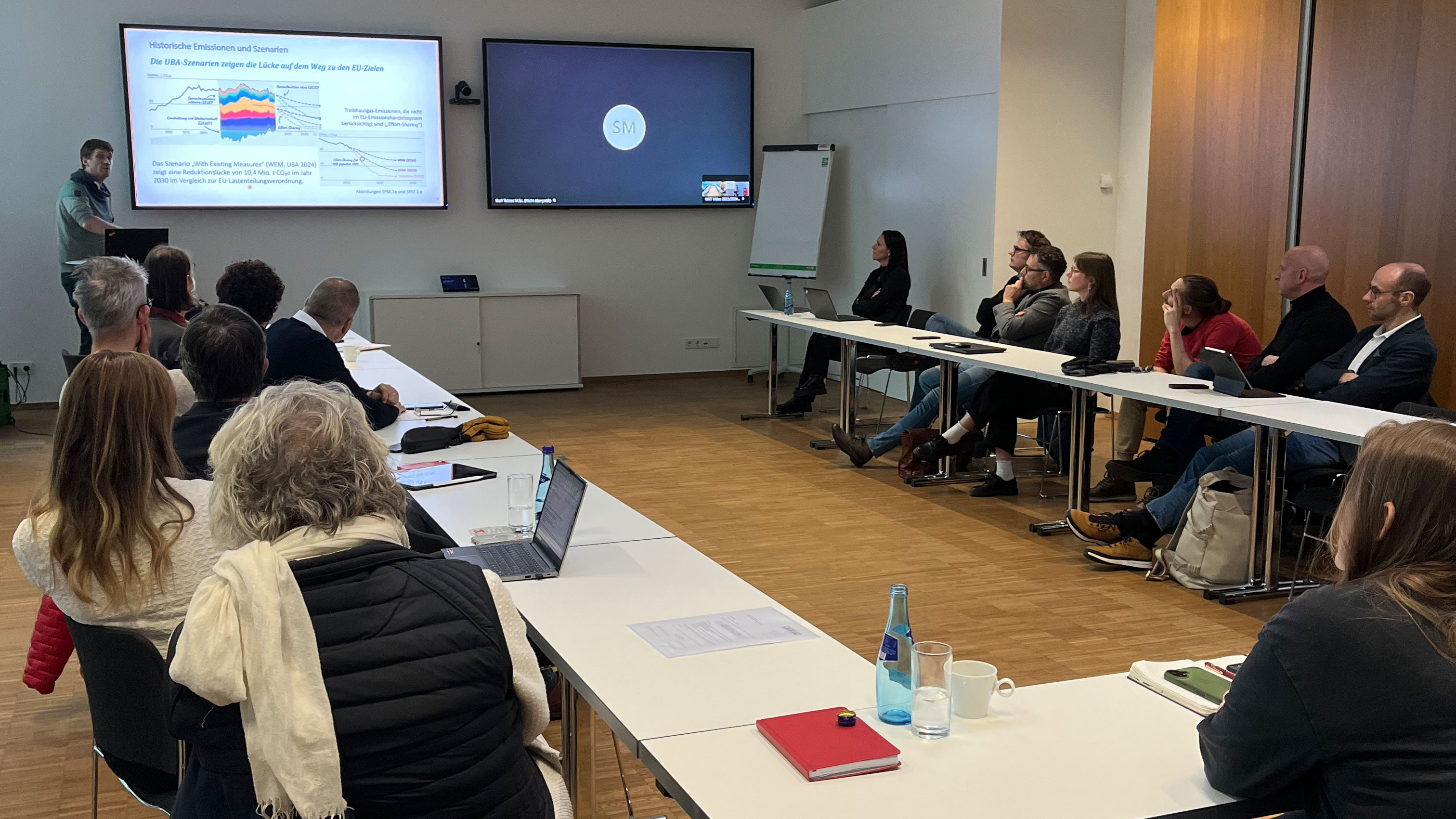 Personen sitzen in einem Seminarraum und h&ouml;ren dem Vortrag des Referenten zu, der vorne steht und seine Folien pr&auml;sentiert