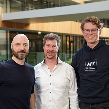 Drei Personen stehen lächelnd nebeneinander in einem modernen, offenen Bürogebäude.