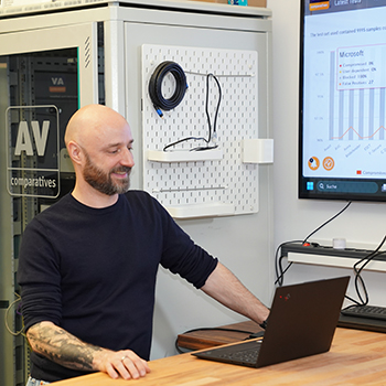 ChatGPT:Person mit rasiertem Kopf arbeitet lächelnd an einem Laptop in einem Technikraum, neben einem Bildschirm mit Präsentationsgrafik.