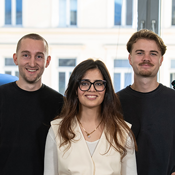 :Drei Personen stehen lächelnd nebeneinander in einem hellen Raum vor großen Fenstern.