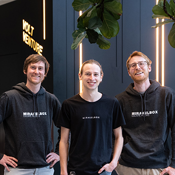 Drei Personen in Mirakelbox-Kleidung stehen lächelnd in einer modernen Eingangshalle vor einer Wand mit der Leuchtschrift „Volt Venture“.