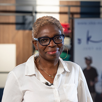 Person mit kurzer, gelockter Frisur und Brille steht lächelnd in einem Bekleidungsgeschäft an einem Tisch mit gefalteter Kleidung.