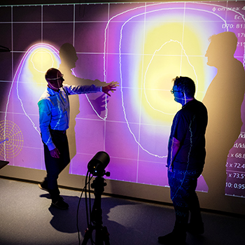 Zwei Personen stehen in einem abgedunkelten Raum vor einer großen, bunt leuchtenden Datenprojektion, eine Person deutet erklärend auf die Grafik