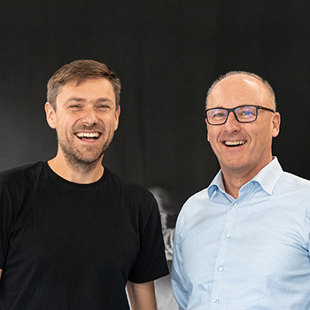 Zwei Personen stehen lächelnd nebeneinander vor einem dunklen Hintergrund.