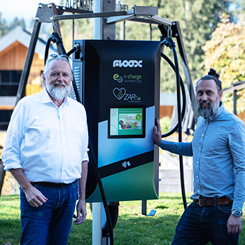 Zwei Personen stehen im Freien neben einer modernen E-Ladesäule, im Hintergrund sind Holzhäuser und Bäume zu sehen.