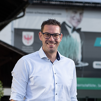 Person mit Brille und weißem Hemd steht lächelnd im Freien vor einer unscharfen Werbetafel.