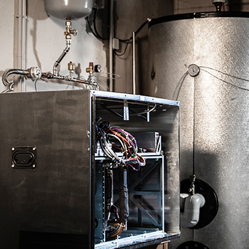 Offener Metallkasten mit sichtbaren Kabeln neben einem großen zylindrischen Heiz- oder Speicherbehälter in einem Technikraum.