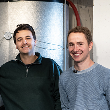 Zwei Personen stehen lächelnd in einem Technikraum neben einem geöffneten Metallkasten mit Kabeln und einem großen silbernen Speicherbehälter.