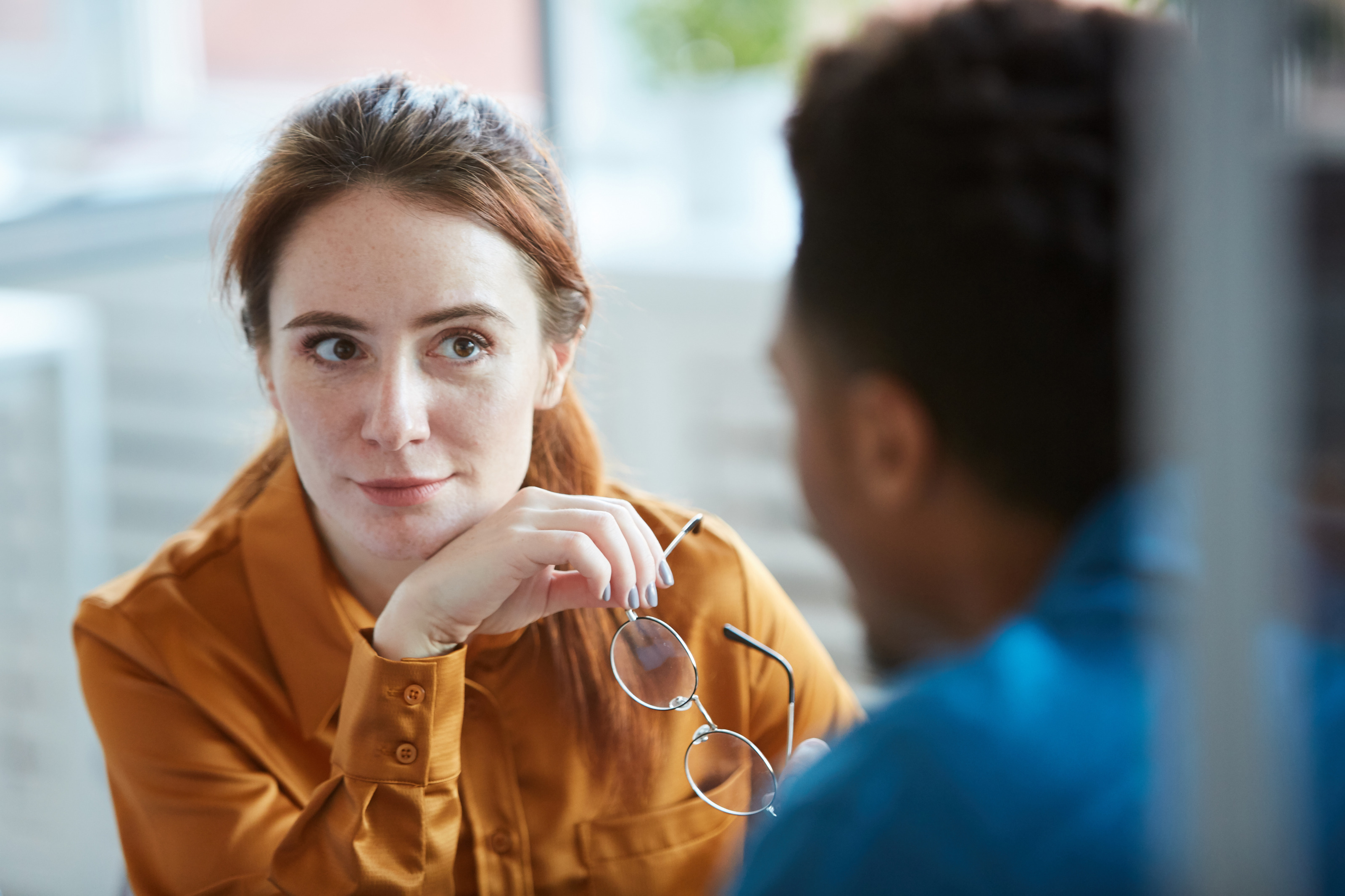 Person im Fokus mit rotbraunen langen Haaren und senfgelber Bluse h&auml;lt Brille in der Hand und blickt interessiert zu einer anderen Person, die in Unsch&auml;rfe dargestellt ist