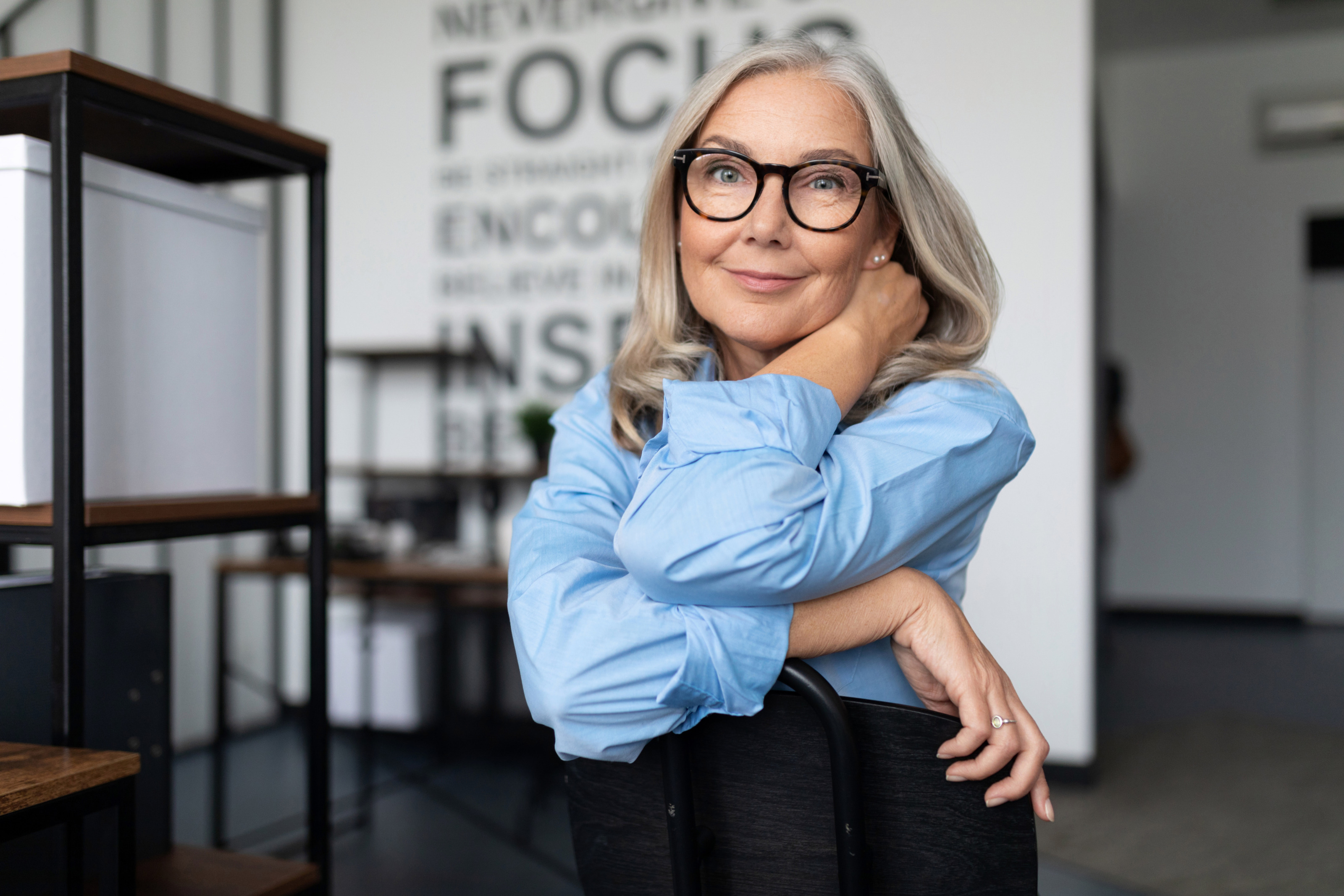 Sanft l&auml;chelnde Peron mit Brillen und wei&szlig;grauem Haar sitzt auf Stuhl und st&uuml;tzt Arme auf R&uuml;ckenlehne, eine Hand an den Hals gelegt, im Hintergrund Regale und wei&szlig;e Wand mit schwarzer Schrift mit Schriftzug Focus