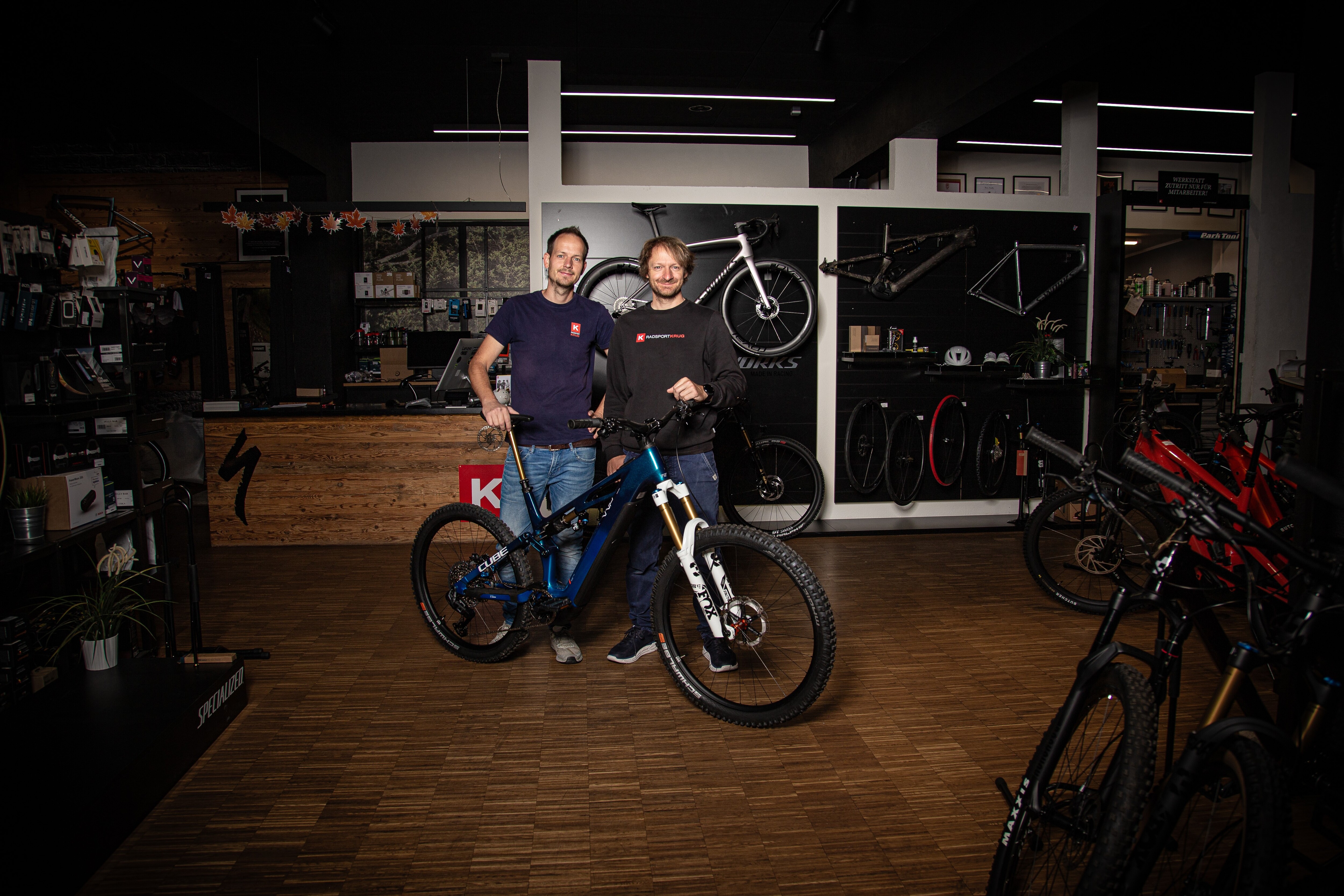 Die Aufnahme zeigt zwei Unternehmer in ihrem Fahrradfachgesch&auml;ft. Sie stehen im Verkaufsraum zwischen mehreren Fahrr&auml;dern und E-Bikes. Hinter ihnen befinden sich an der Wand pr&auml;sentierte Rennr&auml;der, Fahrradrahmen und Zubeh&ouml;r. Rechts ist ein Bereich mit E-Bikes zu sehen, im Hintergrund eine Werkstatt f&uuml;r Service- und Reparaturarbeiten. Die moderne Einrichtung vermittelt den Eindruck eines spezialisierten Fachbetriebs f&uuml;r Verkauf, Wartung und individuelle Beratung im Radsportbereich.