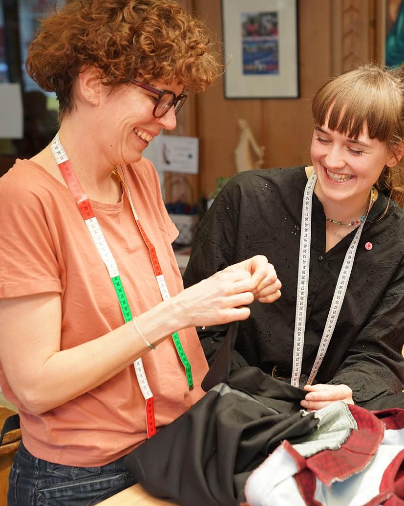 Zwei junge Frauen stehen gemeinsam in einem Raum und betrachten eine Stoffprobe oder N&auml;harbeit.