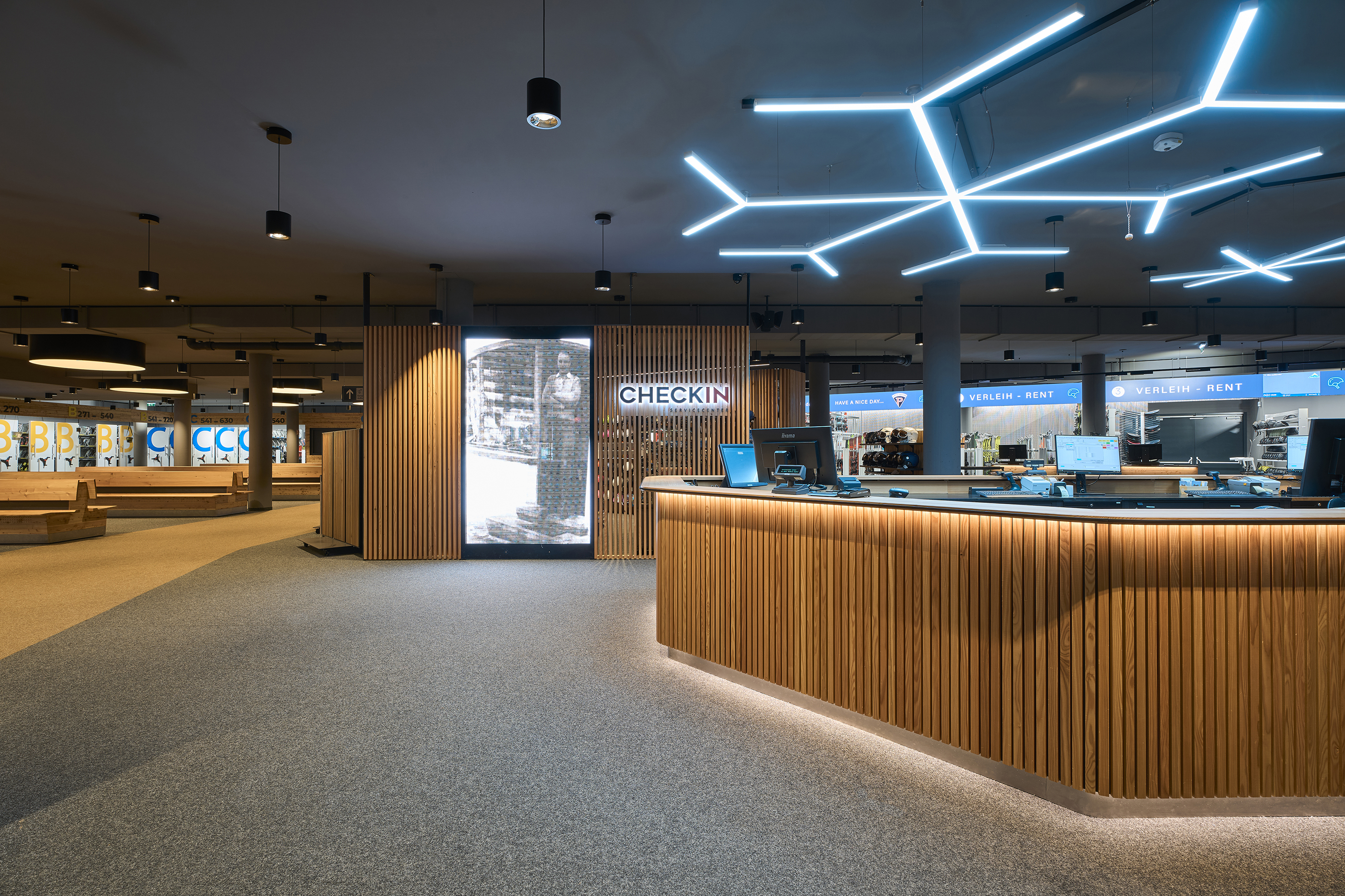 Ein moderner Innenraum mit einem gro&szlig;en Check-in-Tresen aus Holz. &Uuml;ber dem Tresen h&auml;ngen geometrisch angeordnete Leuchtelemente. Im Hintergrund sind weitere Servicebereiche und Beschilderungen zu sehen.