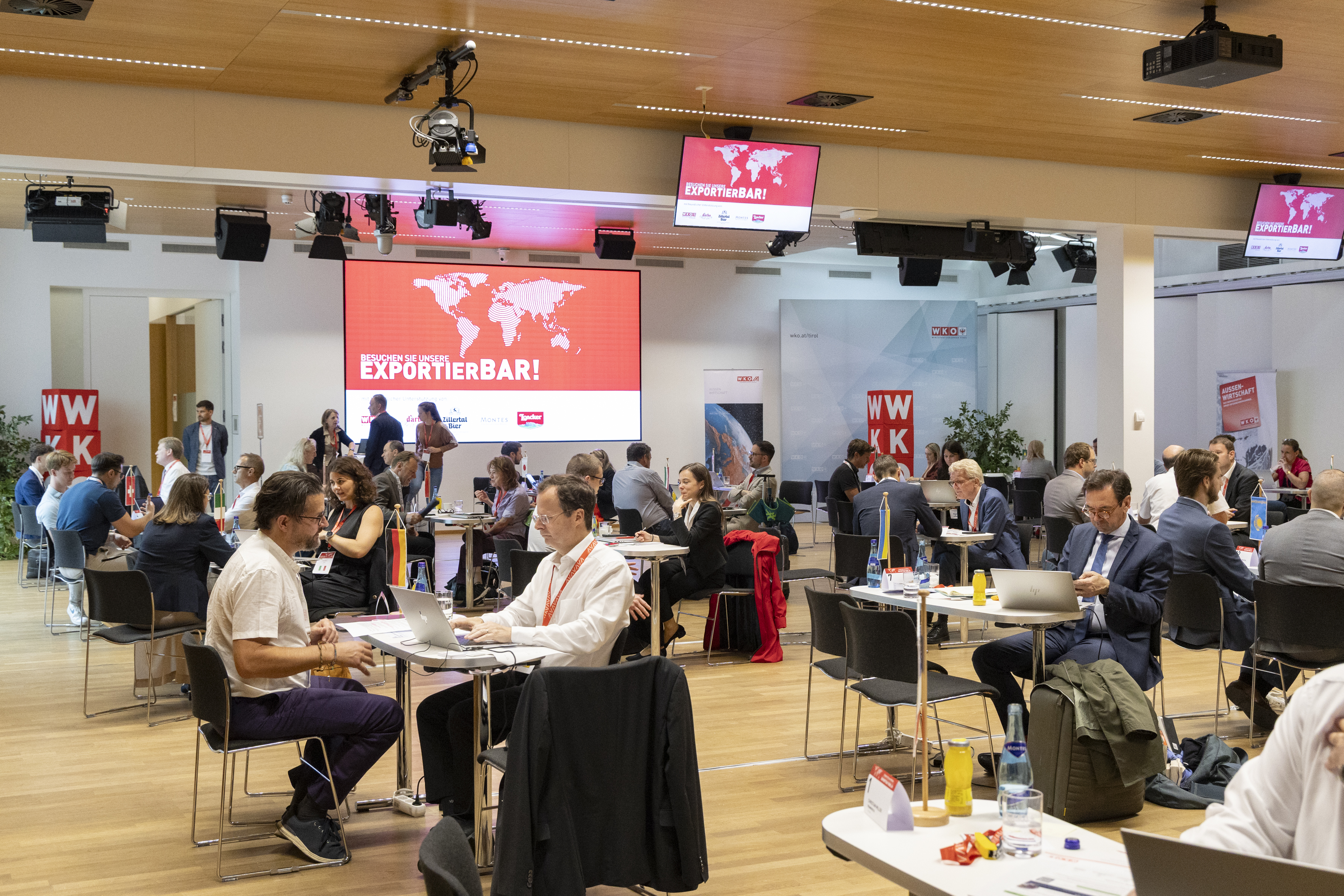 Mehrere Personen sitzen an Tischen in einem Veranstaltungsraum und f&uuml;hren Gespr&auml;che. Im Hintergrund ist eine gro&szlig;e Leinwand mit der Aufschrift &bdquo;Besuchen Sie unsere Exportierbar!&ldquo; zu sehen.