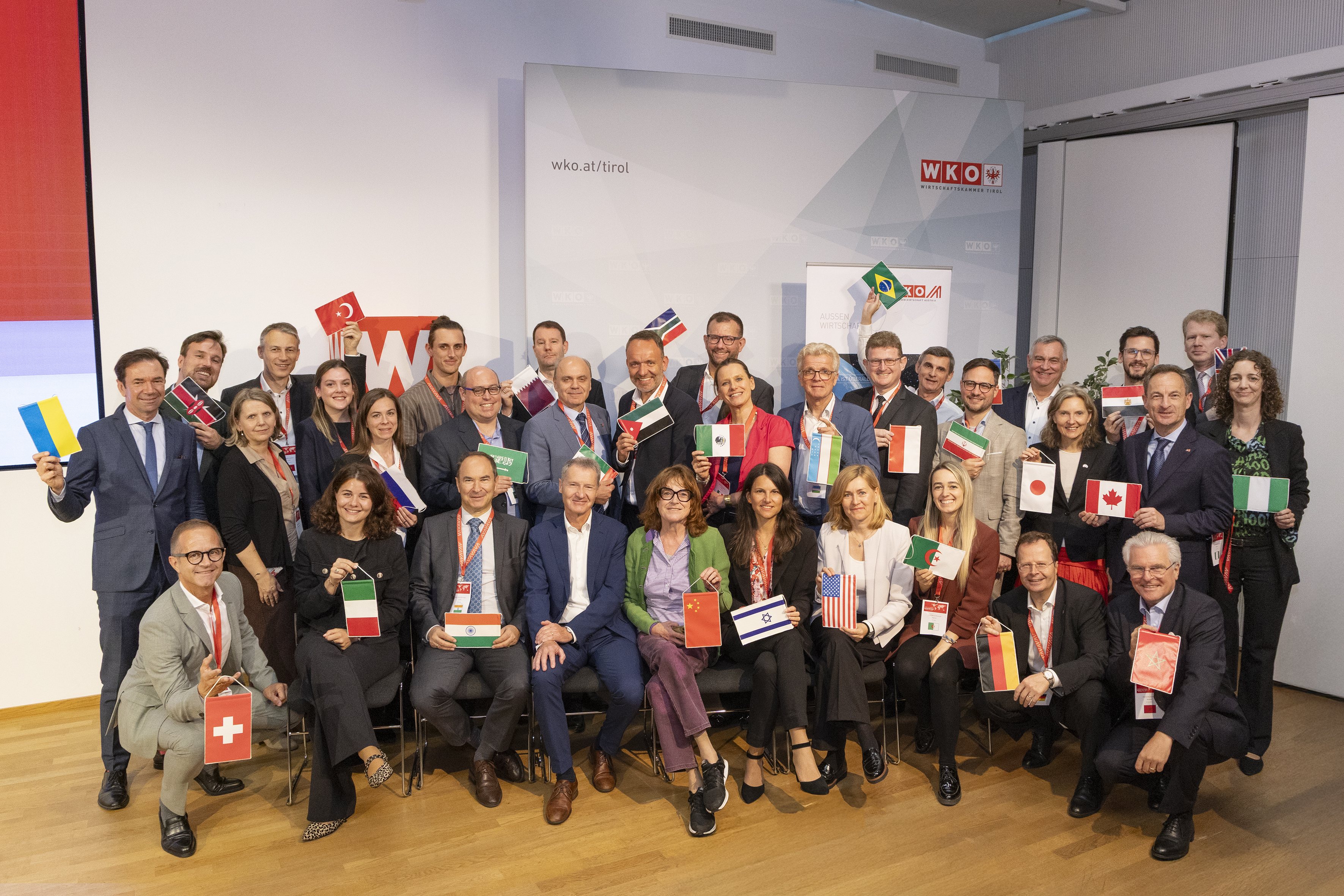 Rund 30 Personen posieren in einem Veranstaltungsraum f&uuml;r ein Gruppenfoto und halten kleine L&auml;nderflaggen in die Kamera. Im Hintergrund sind Elemente der Wirtschaftskammer Tirol sichtbar.