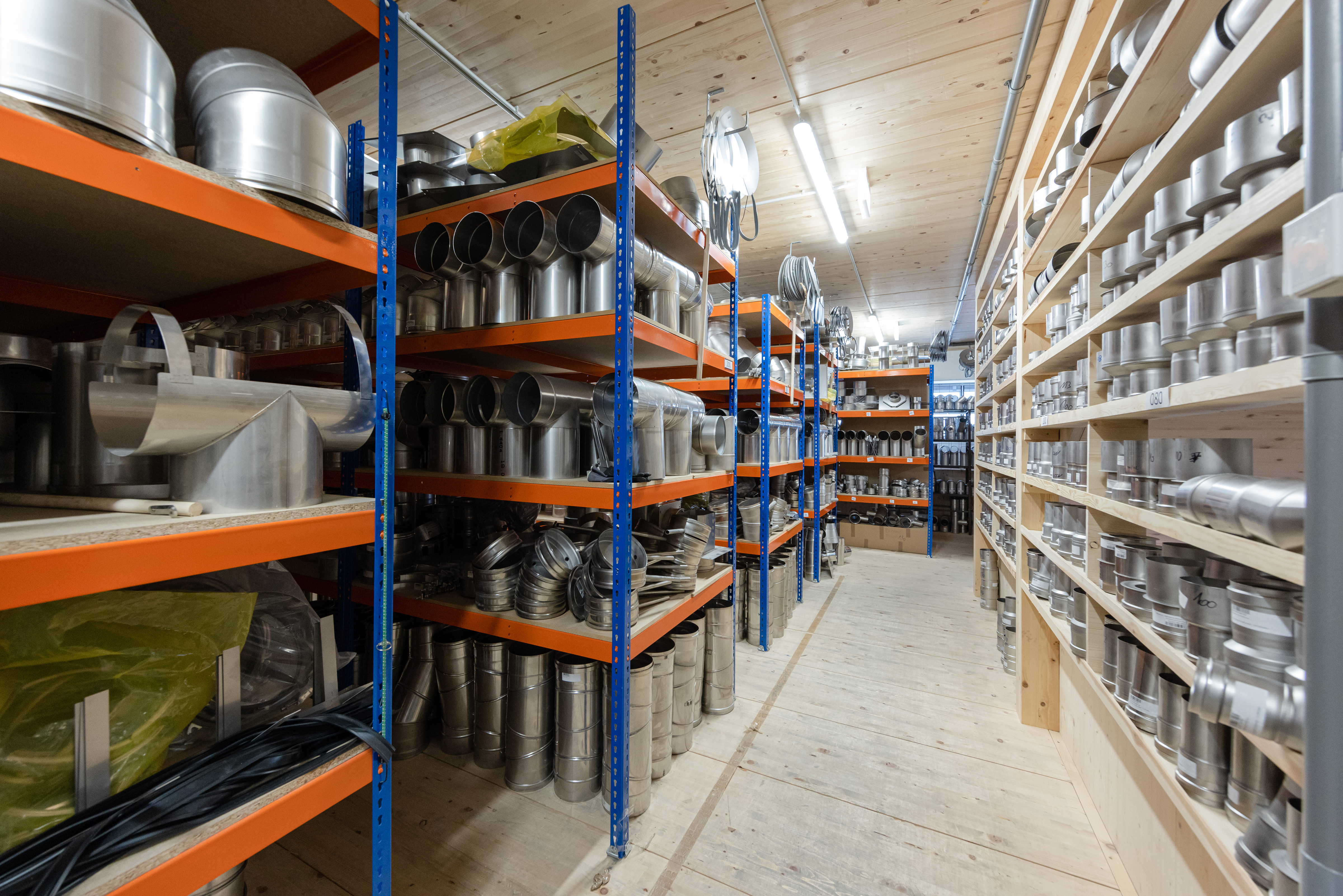 Ein gro&szlig;es Lager mit langen Regalen voller Edelstahlteile f&uuml;r Kamin- und Abgassysteme. Auf beiden Seiten des Gangs stehen ordentlich sortierte Metallrohre, Verbindungsst&uuml;cke und Zubeh&ouml;r. Der Raum ist hell beleuchtet und vermittelt den Eindruck eines gut organisierten Familienbetriebs mit gro&szlig;em Lagerbestand.