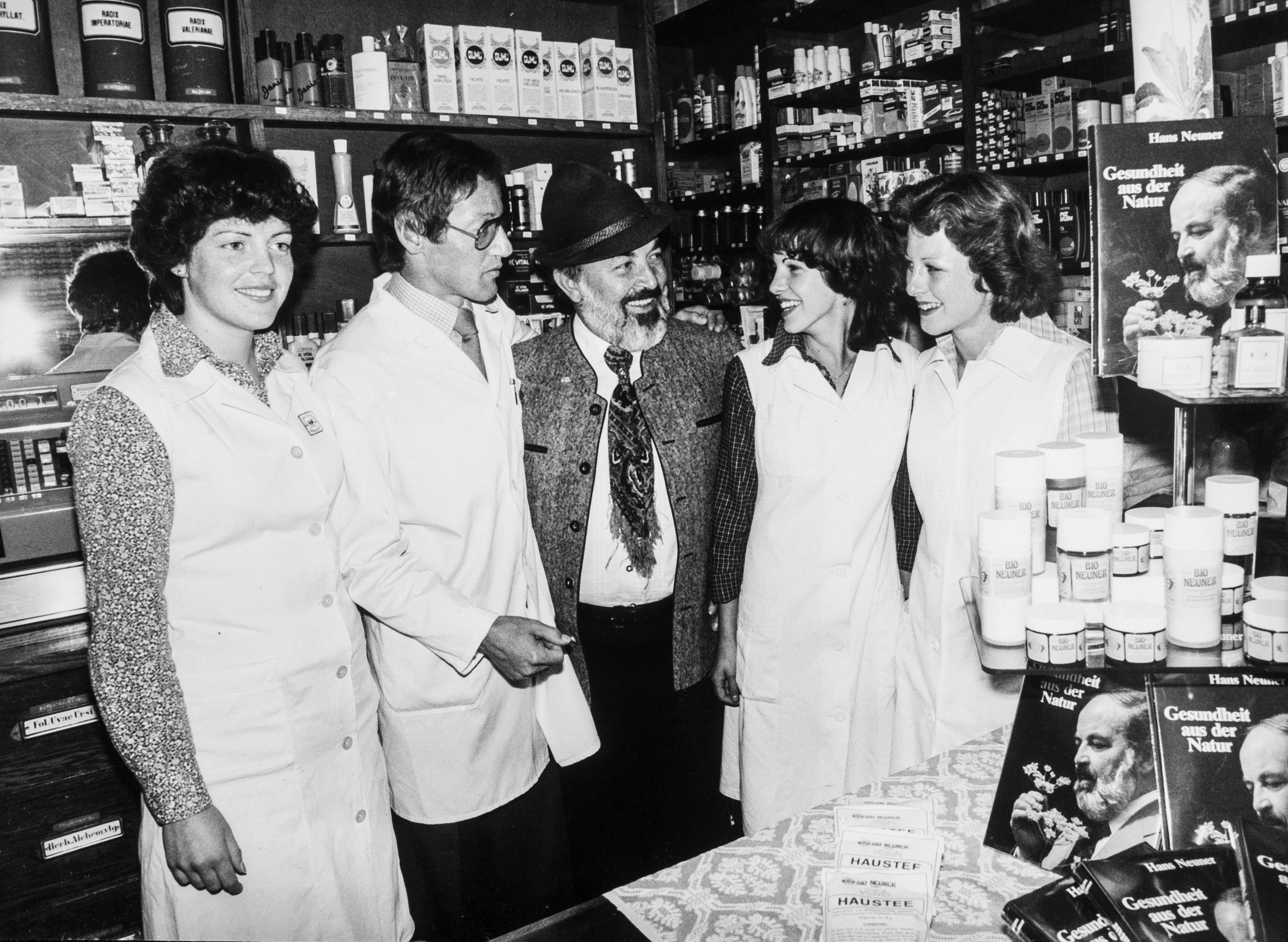Historisches Schwarz-Wei&szlig;-Foto in einer Drogerie mit einem b&auml;rtigen Mann und mehreren Mitarbeiterinnen und Mitarbeitern zwischen Regalen mit Naturprodukten.