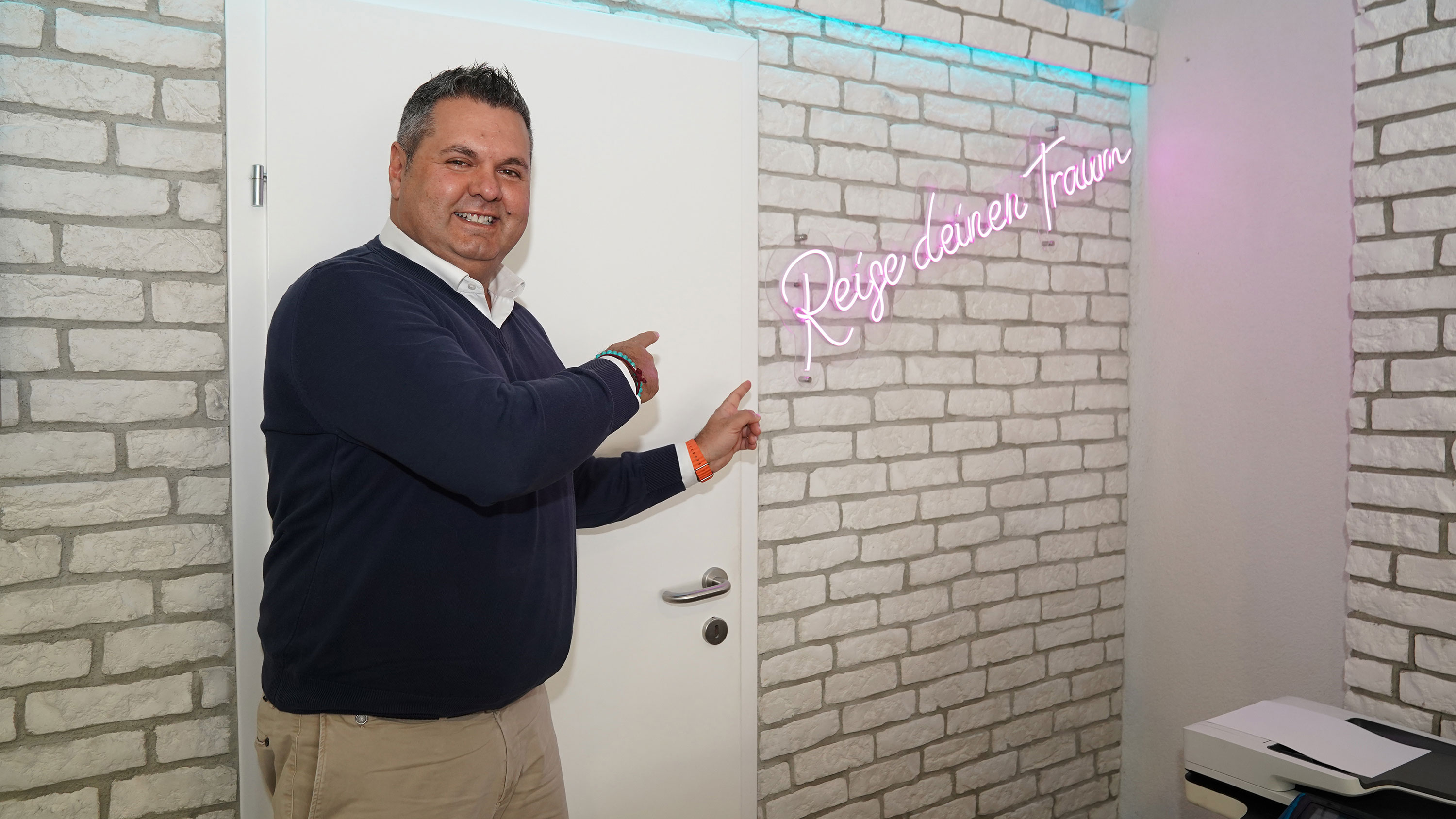 Bora Eker zeigt auf ein Leuchtschild, das an der Wand h&auml;ngt. Darauf steht Reise deinen Traum