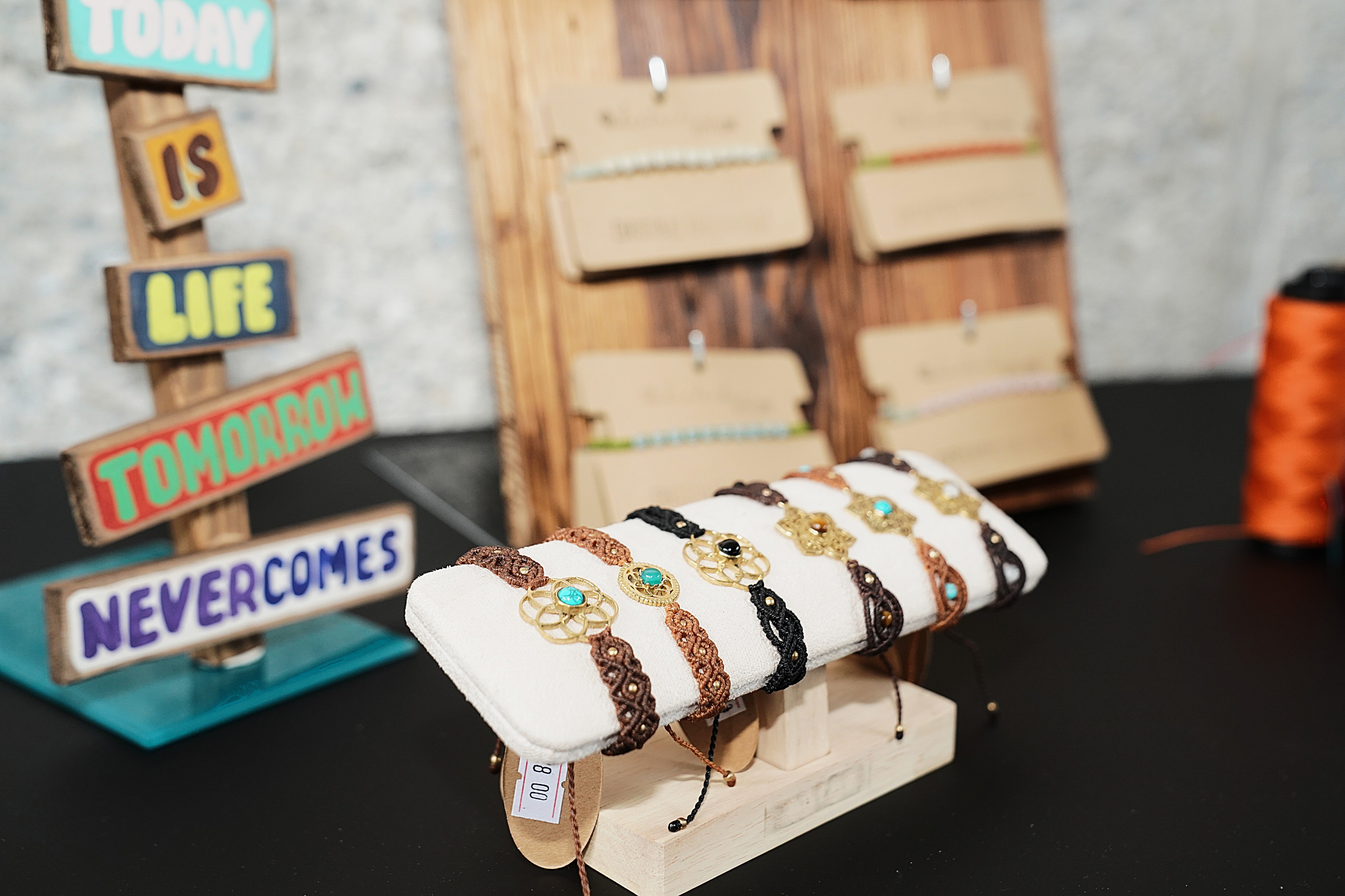 Mehrere handgekn&uuml;pfte Armb&auml;nder liegen auf einem Schmuckst&auml;nder; dahinter stehen weitere Schmuckst&uuml;cke und Garnrollen.