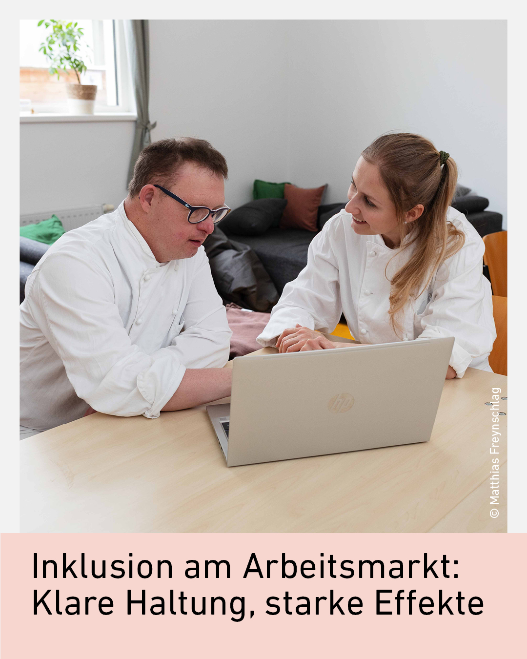 Zwei Personen in wei&szlig;en Arbeitsjacken sitzen an einem Tisch mit einem Laptop und unterhalten sich.
