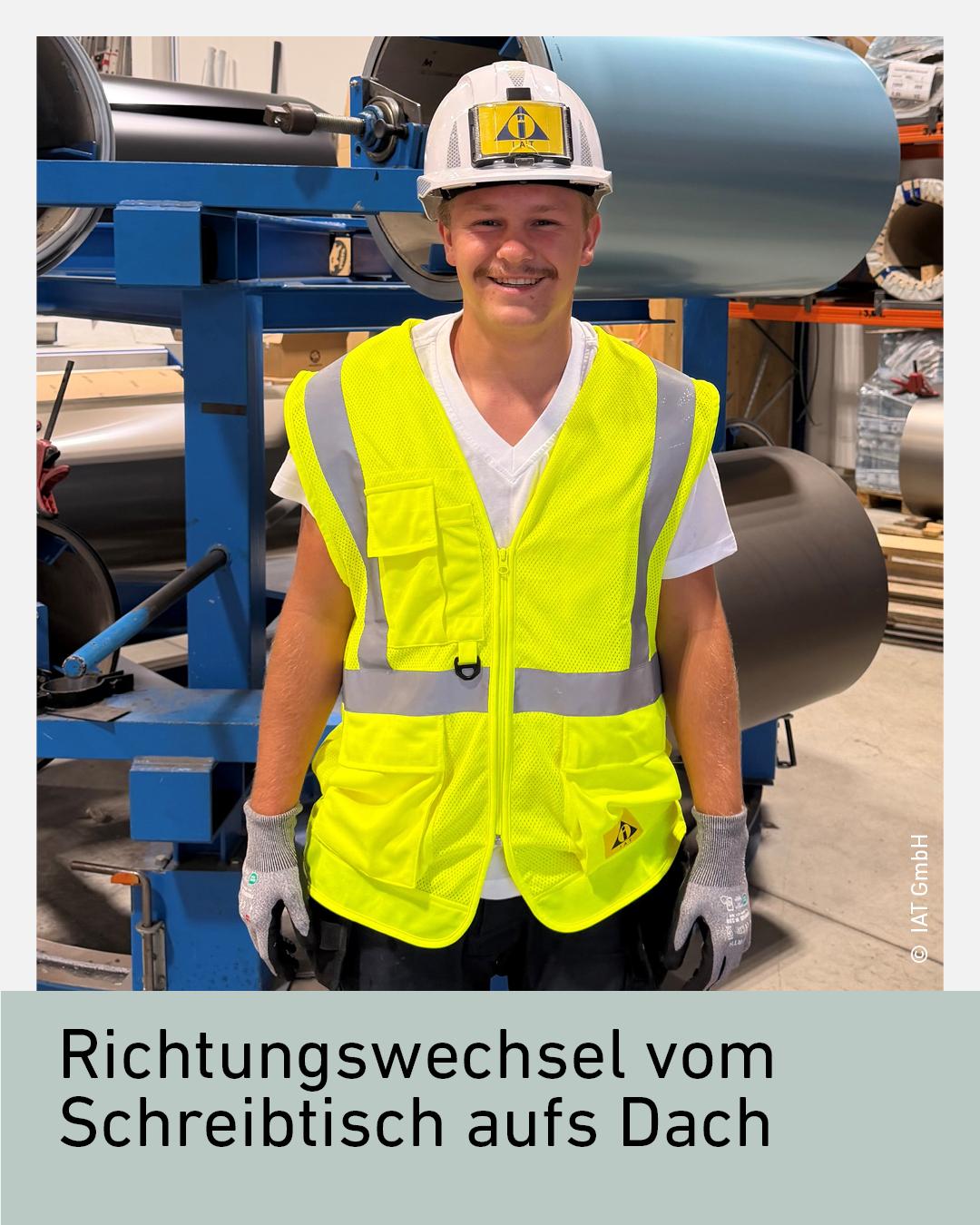 Ein junger Mann in einer gelben Sicherheitsweste und einem Schutzhelm steht vor einer Maschine in einer Industrieumgebung.