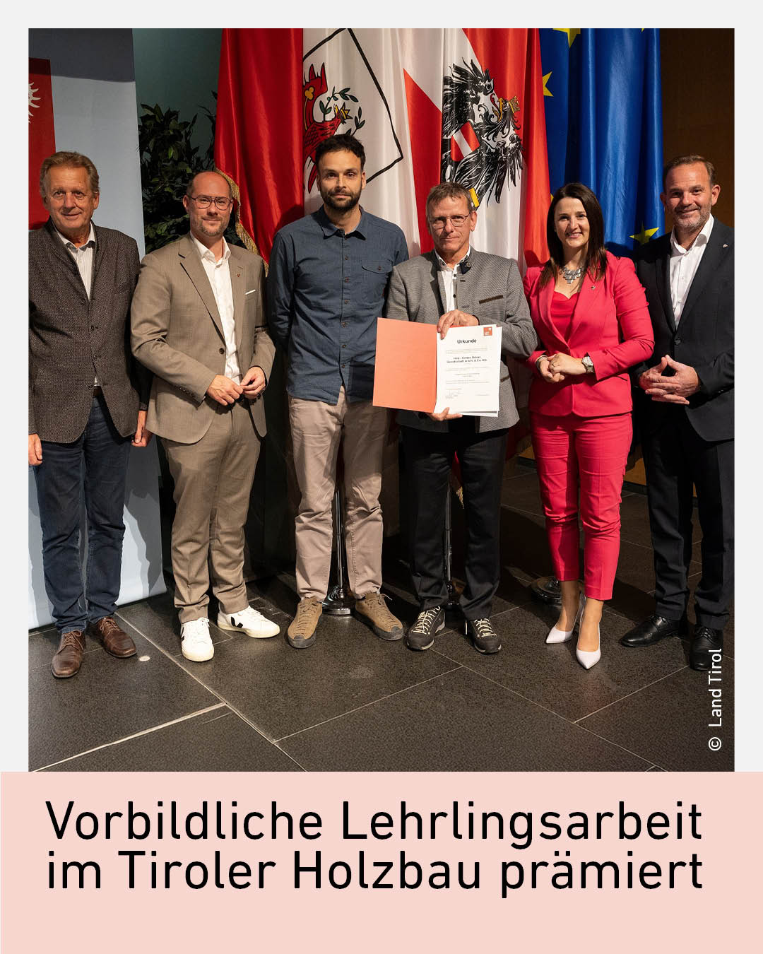 Gruppenfoto bei der Pr&auml;mierung vorbildlicher Lehrlingsarbeit im Tiroler Holzbau mit Vertreterinnen und Vertretern aus Wirtschaft und Politik; &Uuml;bergabe einer Urkunde vor Tiroler und EU Flaggen im Rahmen einer offiziellen Auszeichnung.
