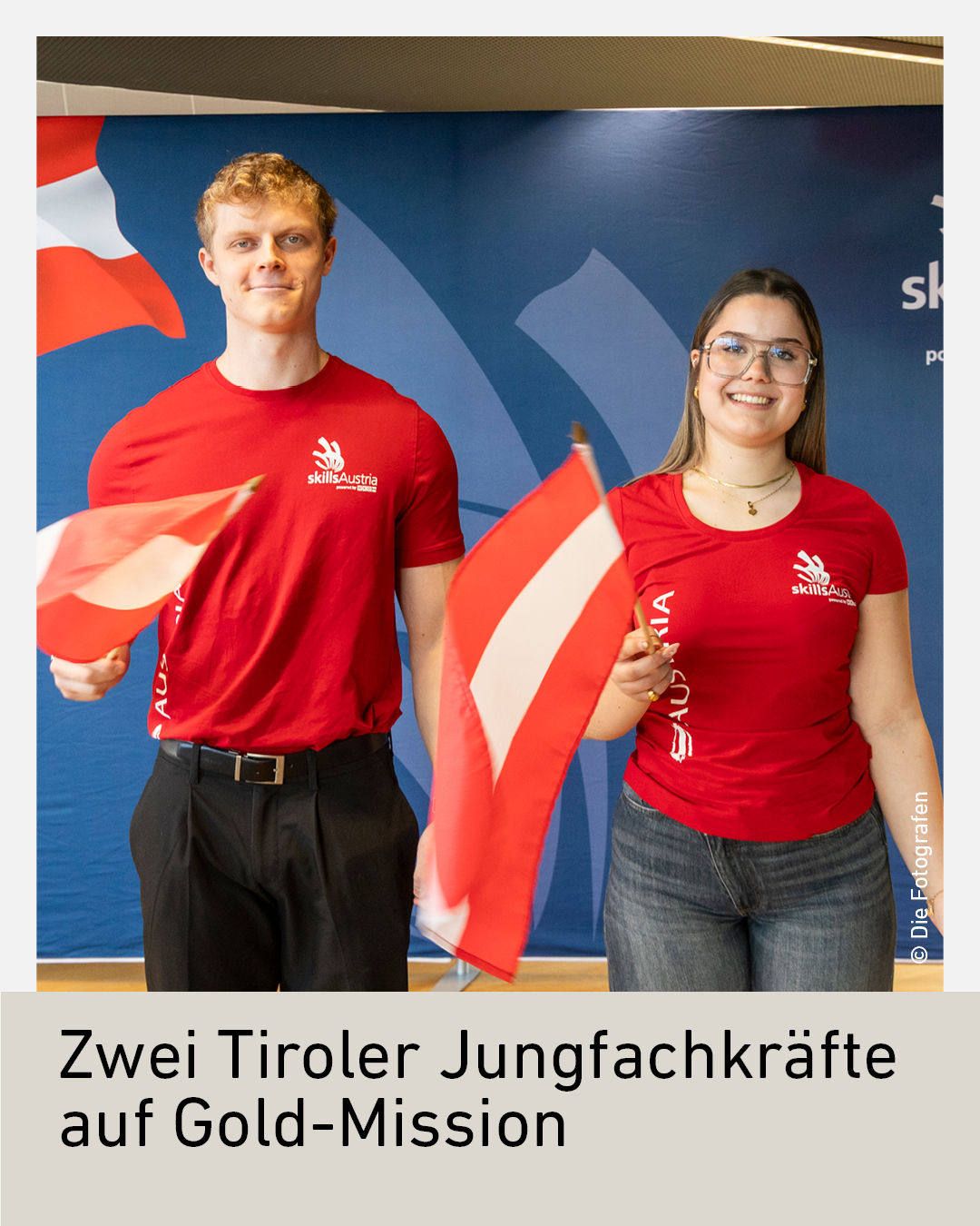 Two people in red shirts holding Austrian flags stand in front of a backdrop with the Skills Austria logo.