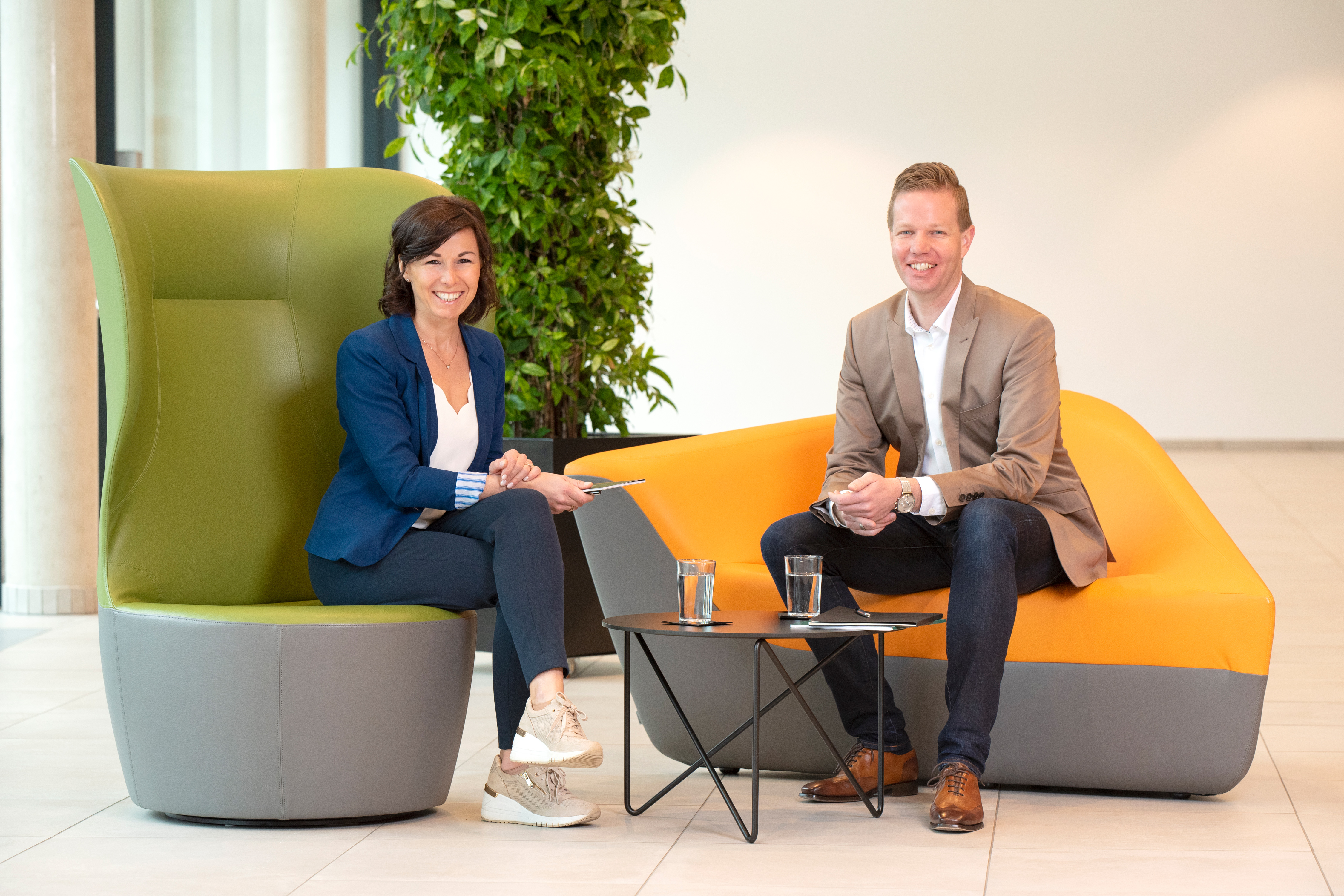 Zwei Personen sitzen in einem modernen, hellen Innenraum auf farbigen Designer-Sesseln. Beide lächeln und halten Notizen in der Hand. Zwischen ihnen steht ein kleiner schwarzer Tisch mit zwei Gläsern Wasser. Im Hintergrund befindet sich eine große grüne Pflanze.