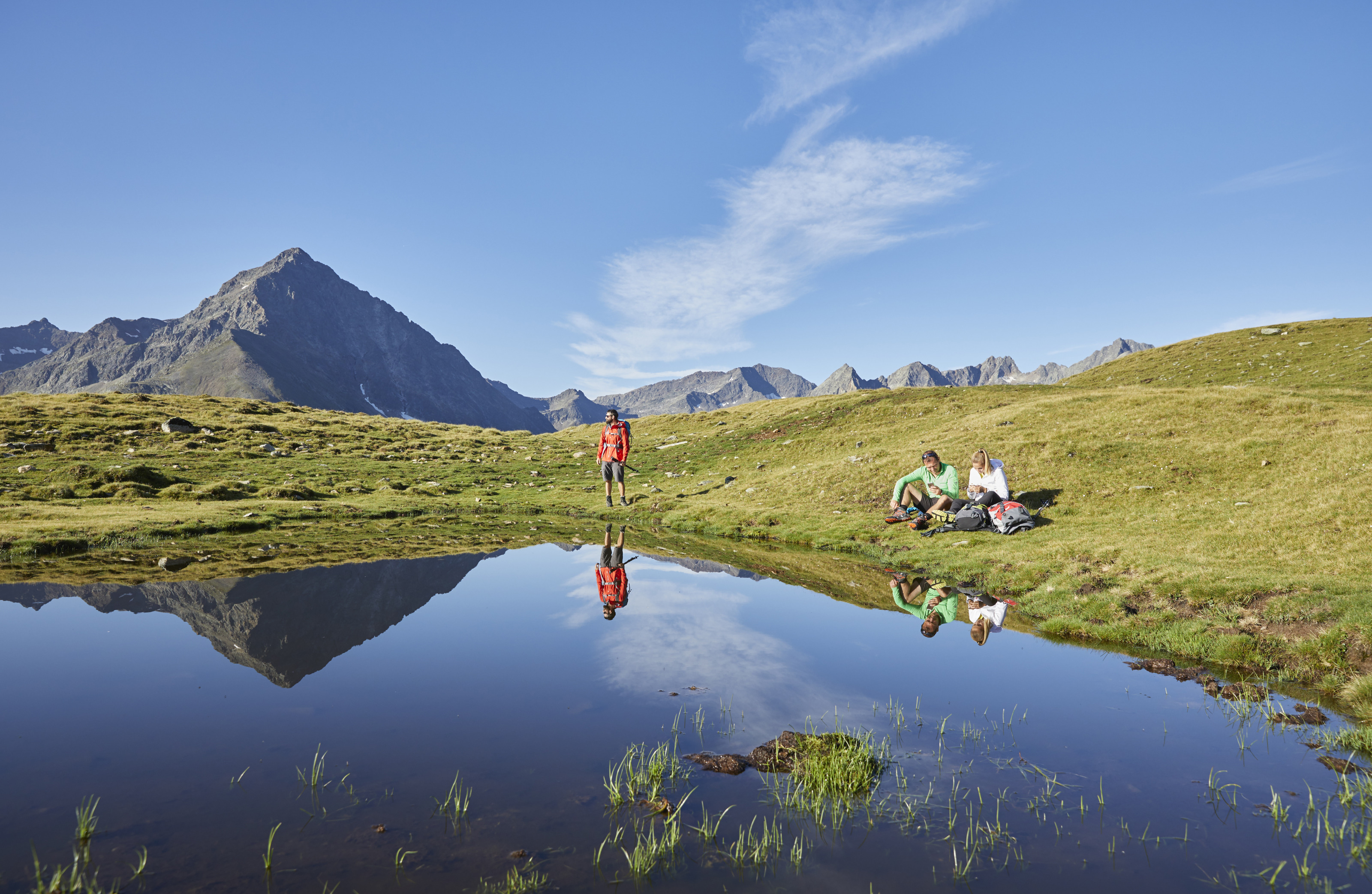 Zwei Personen in Wiese an Bergsee sitzend, eine weitere anbei stehend, spiegeln sich in Gewässer, im Hintergrund Bergkette und blauer Himmel