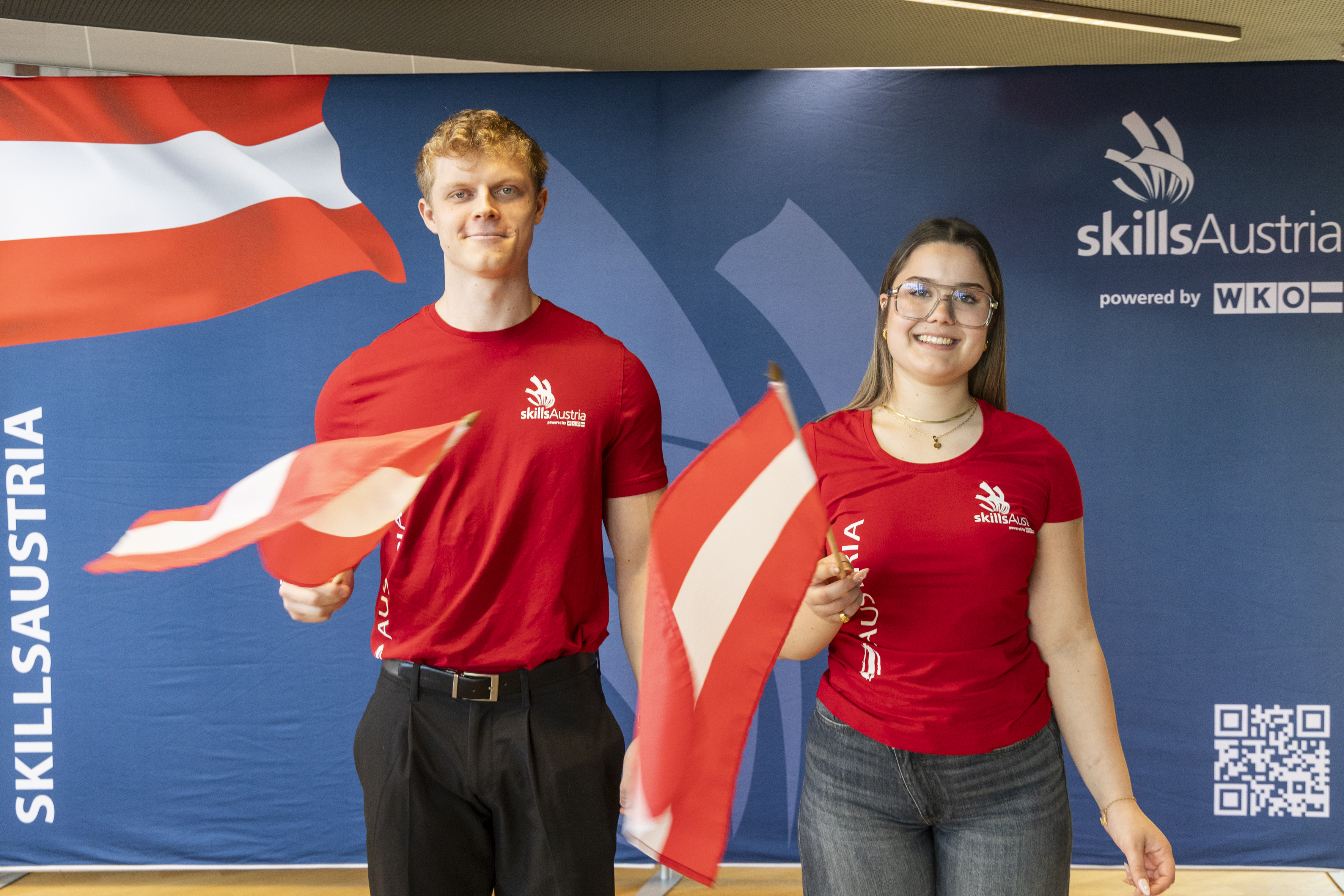 Zwei Tiroler Teilnehmer:innen der WorldSkills 2026 stehen vor einer SkillsAustria-Wand und schwenken &Ouml;sterreich-Flaggen. Beide tragen rote Teamshirts von SkillsAustria.