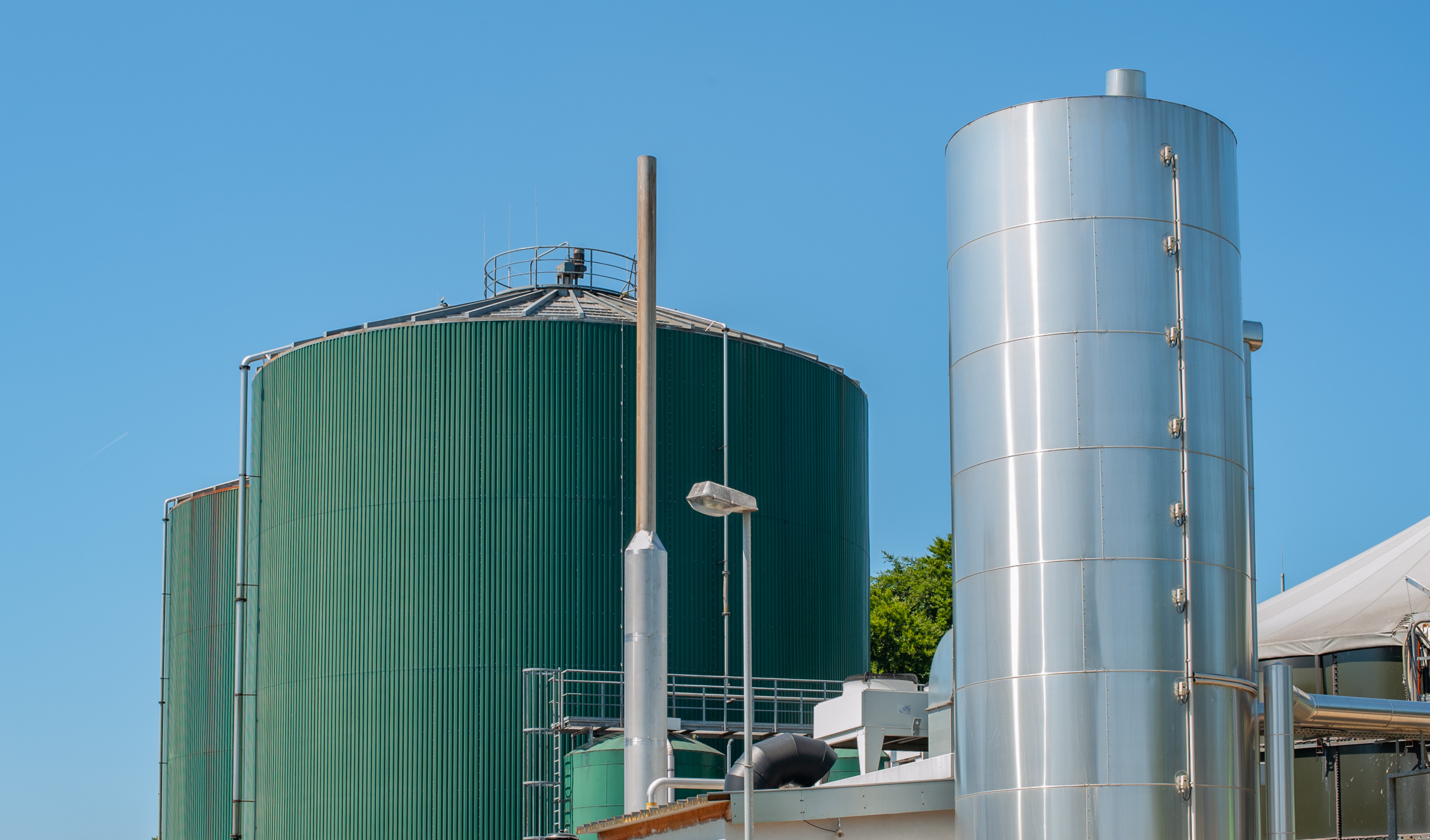 Au&szlig;enansicht einer Biogasanlage: Gro&szlig;e gr&uuml;ne Metallsilos neben silbernen Metallturm unter blauem Himmel