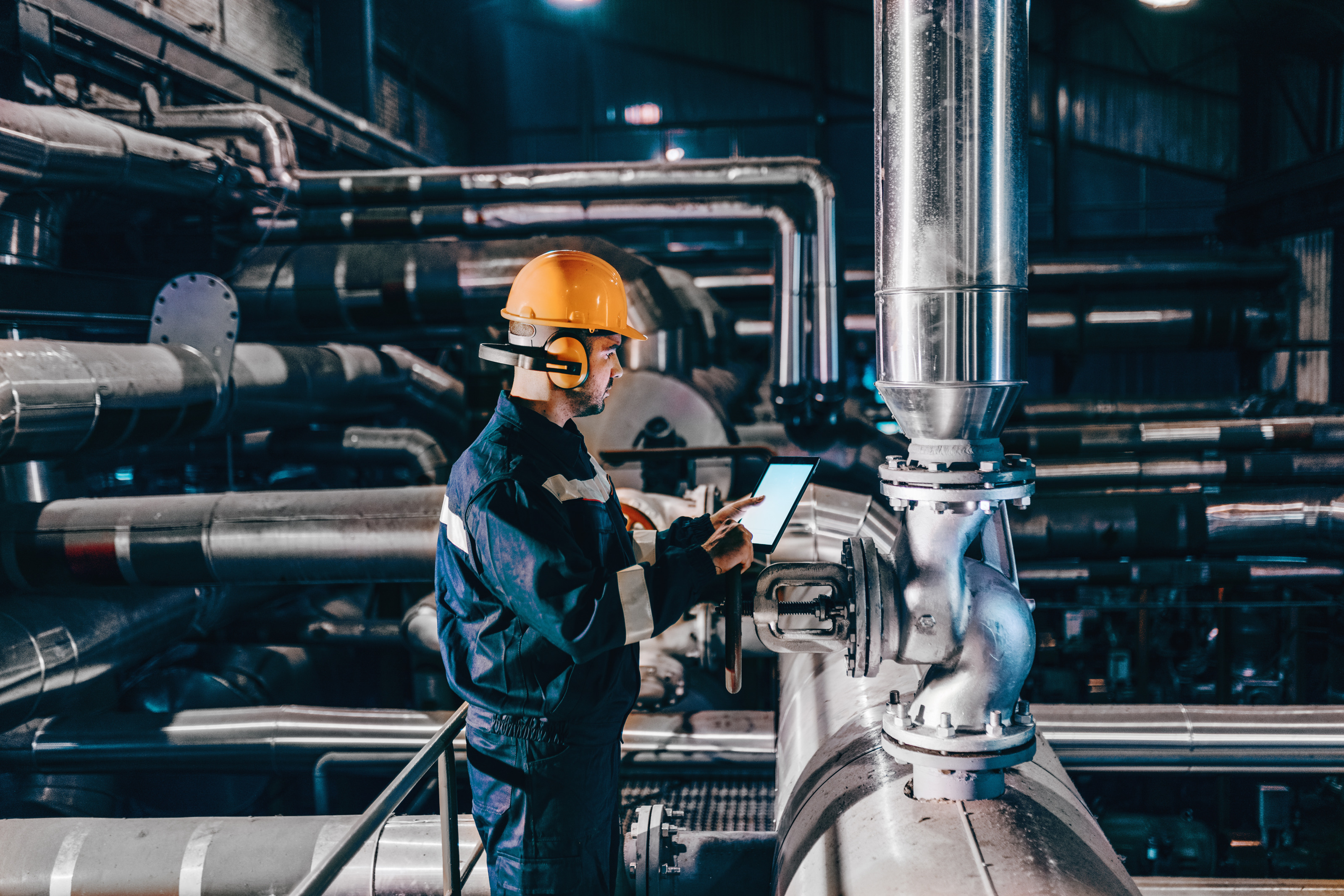 Person in Arbeitskleidung mit orangem Helm und Geh&ouml;rschutz in Seitenansicht steht in Industrieanlage vor gro&szlig;en Metallrohren und h&auml;lt Tablet in H&auml;nden