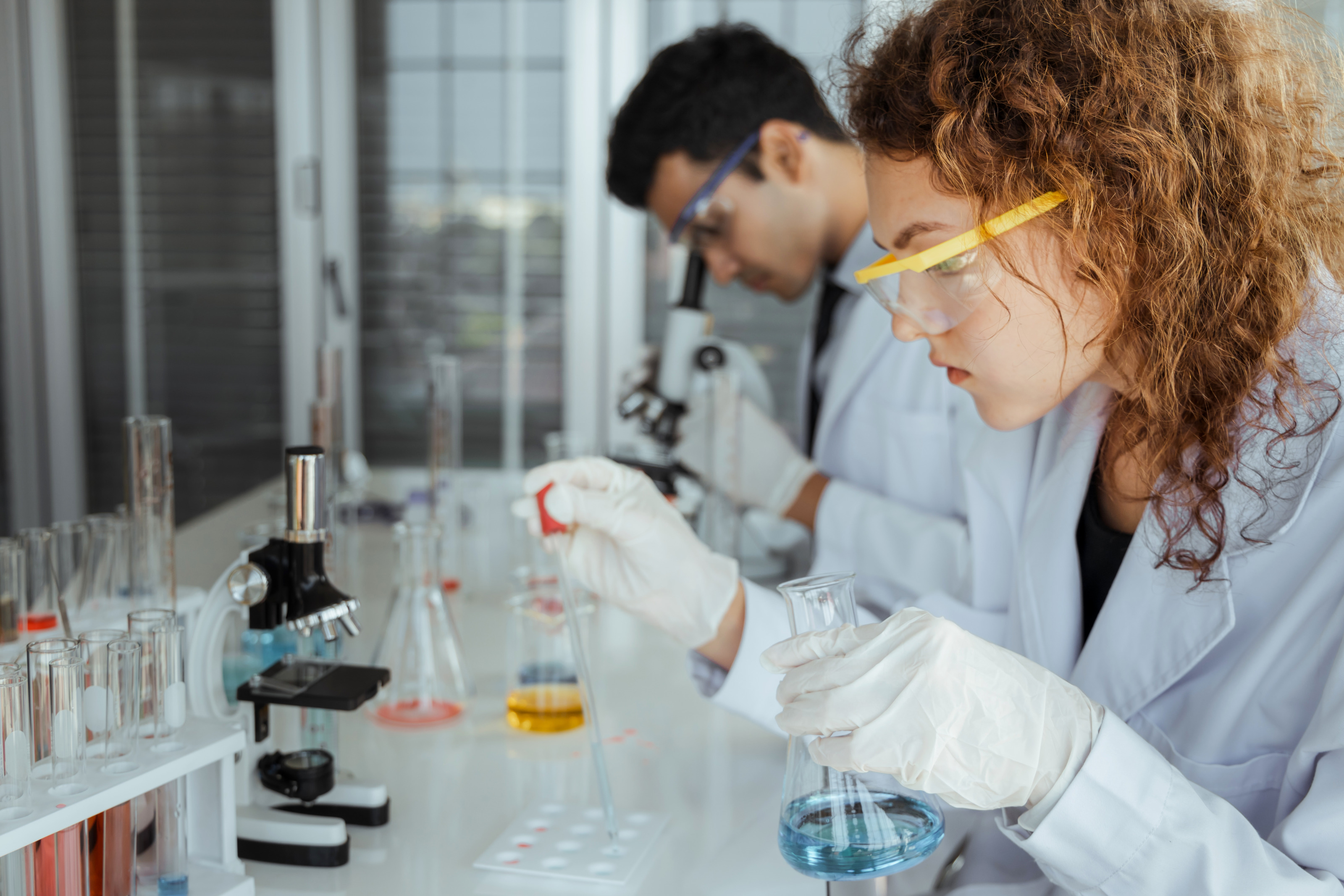 Person mit Schutzbrillen in wei&szlig;em Kittel an Tisch sitzend, mit Pipette Fl&uuml;ssigkeit in Sch&auml;lchen tropfend h&auml;lt in Hand Glas mit Fl&uuml;ssigkeit, am Tisch Mikroskop und R&ouml;hrchen mit Fl&uuml;ssigkeiten, im Hintergrund weitere Person durch Mikroskop &auml;ugend