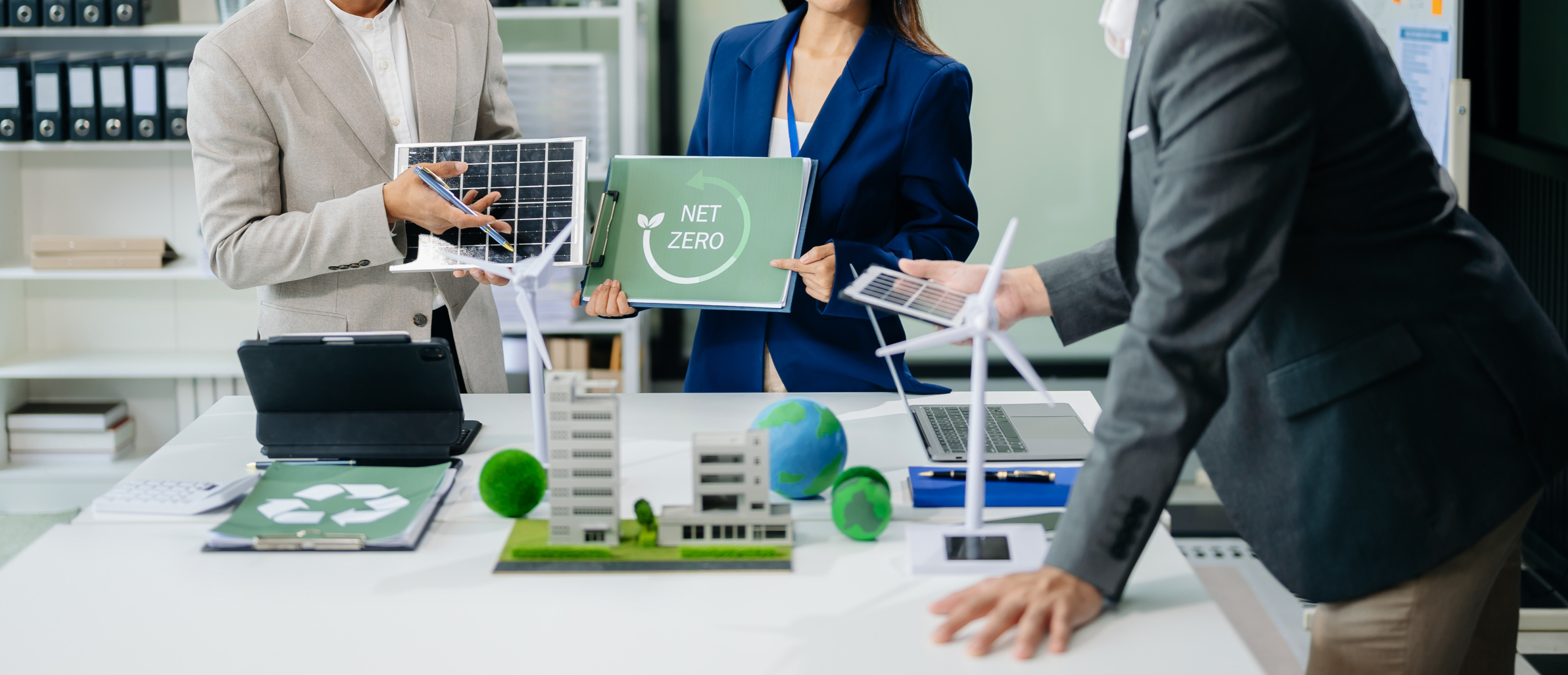 Mehrere Personen im Ausschnitt, nur Oberk&ouml;rper und H&auml;nde ersichtlich, an Tisch stehend mit Modellen wie das eines Windrades, eine Person h&auml;lt Schild mit Aufschrift Net Zero in H&auml;nden