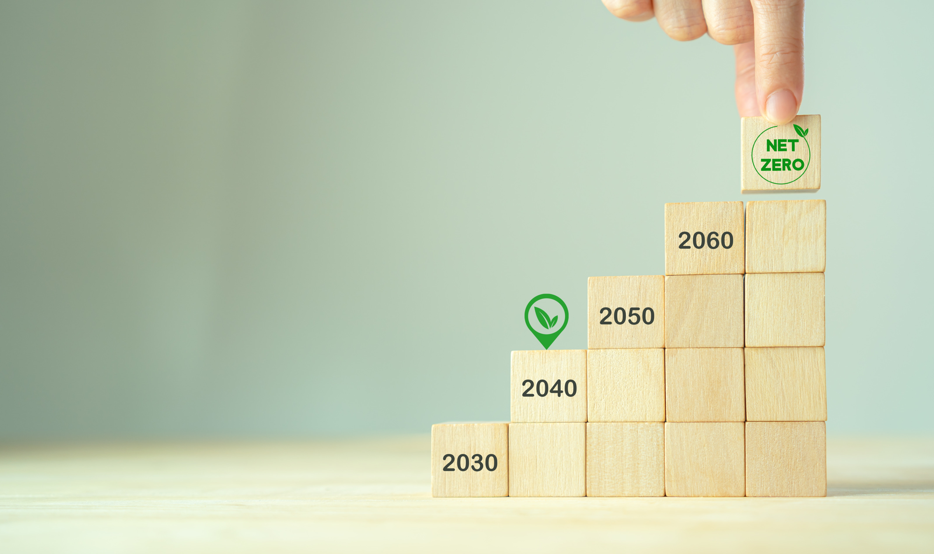 Holzw&uuml;rfel neben- und aufeinander gereiht aufsteigende Stufen formend, die ersten vier Stufen mit den Aufschriften 2030,2040,2050 und 2060, der oberste W&uuml;rfel mit gr&uuml;ner Aufschrift Net Zero wird von einer Hand platziert