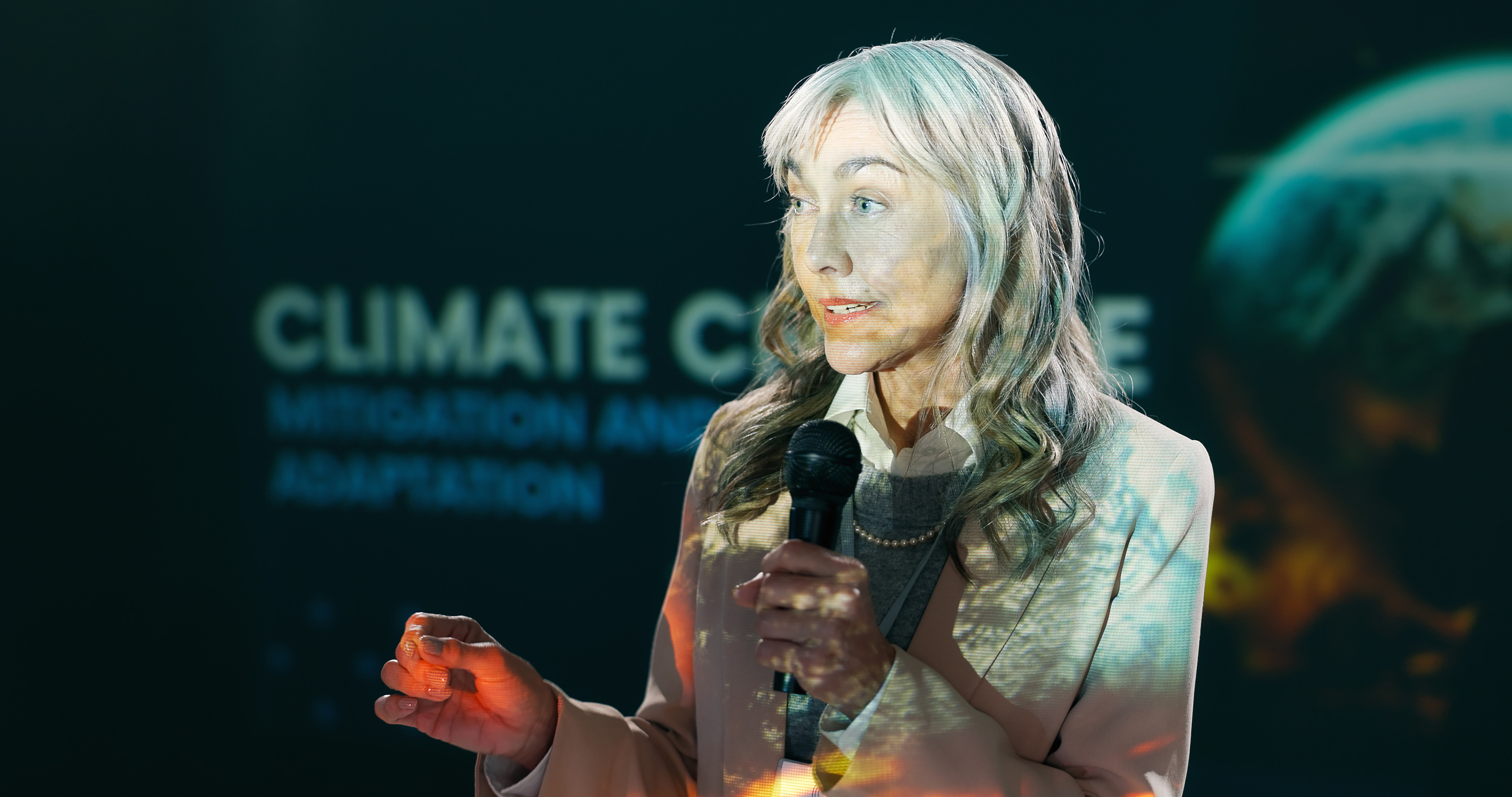 Eine Person mit offenem Haar, hellem Sakko und Mikrofon in der linken Hand steht vor einem gro&szlig;en Bildschirm und spricht in das Mikrofon. Auf dem Bildschirm steht Climate Change
